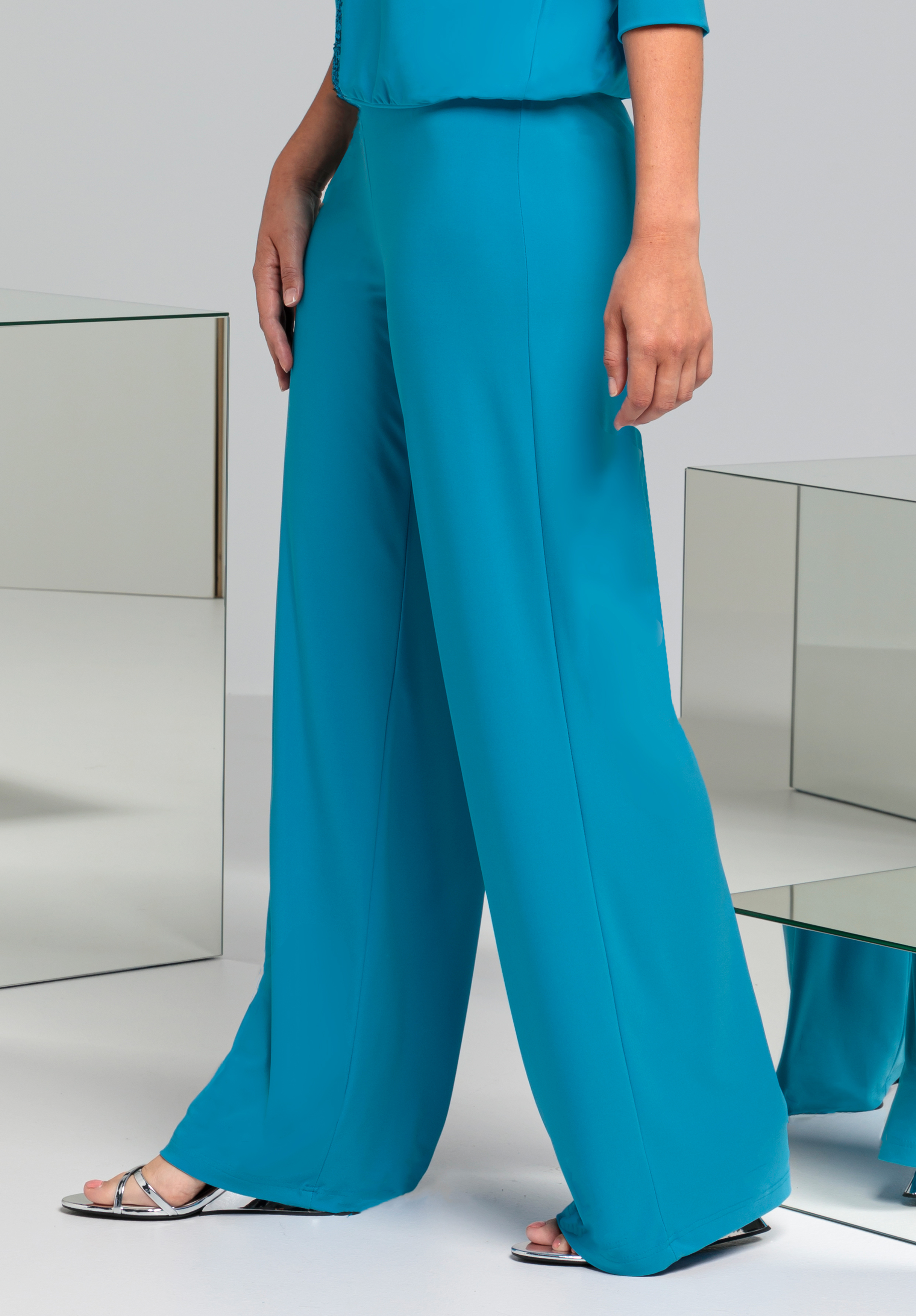 Eine Person steht in einer lockeren, leuchtend türkisfarbenen Hose und einem dazu passenden Oberteil. Sie trägt silberne Pumps mit offener Spitze. Im Hintergrund spiegeln sich die Oberflächen, der Boden ist hellgrau.