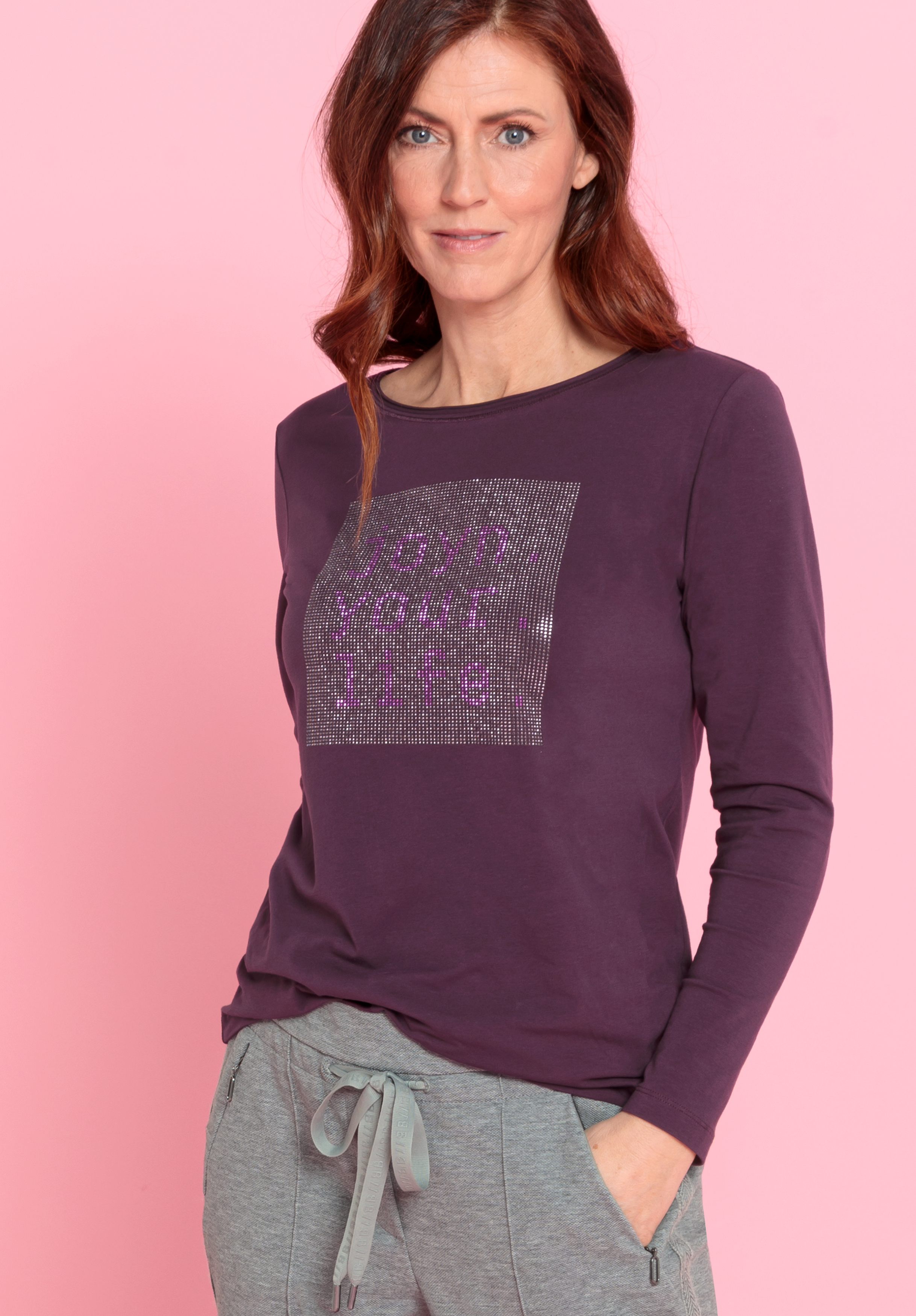 Rundhalsshirt DAMI Eine Frau mit langen braunen Haaren trägt ein lilafarbenes, langärmeliges Oberteil mit einem glitzernden, quadratischen Muster und eine graue Hose mit Kordelzug. Sie steht vor einem rosafarbenen Hintergrund und hat eine Hand in ihrer Tasche.