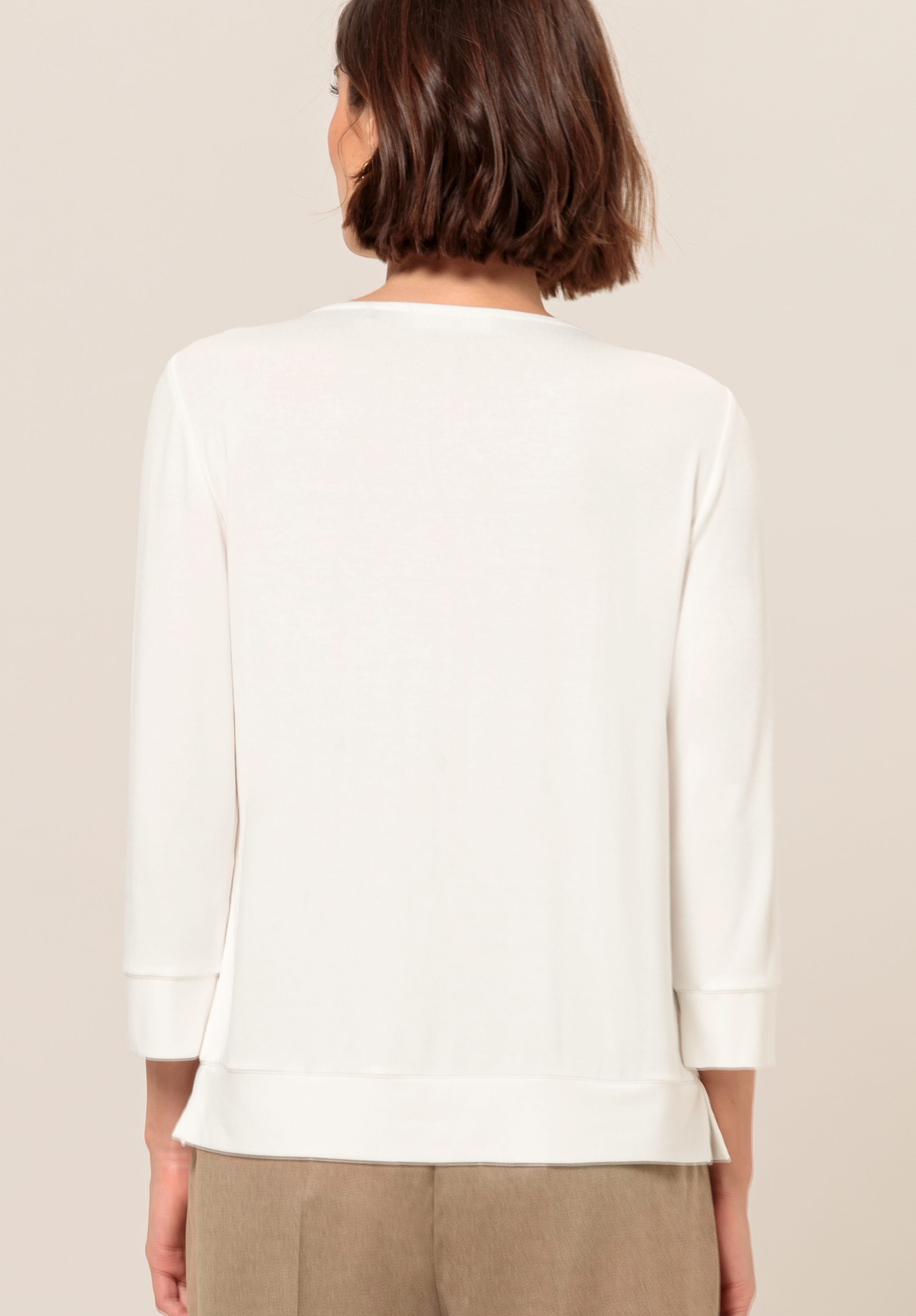 Eine Frau mit kurzen braunen Haaren ist von hinten zu sehen. Sie trägt ein langärmliges, cremefarbenes Oberteil und hellbraune Hosen vor einem schlichten, hellen Hintergrund.