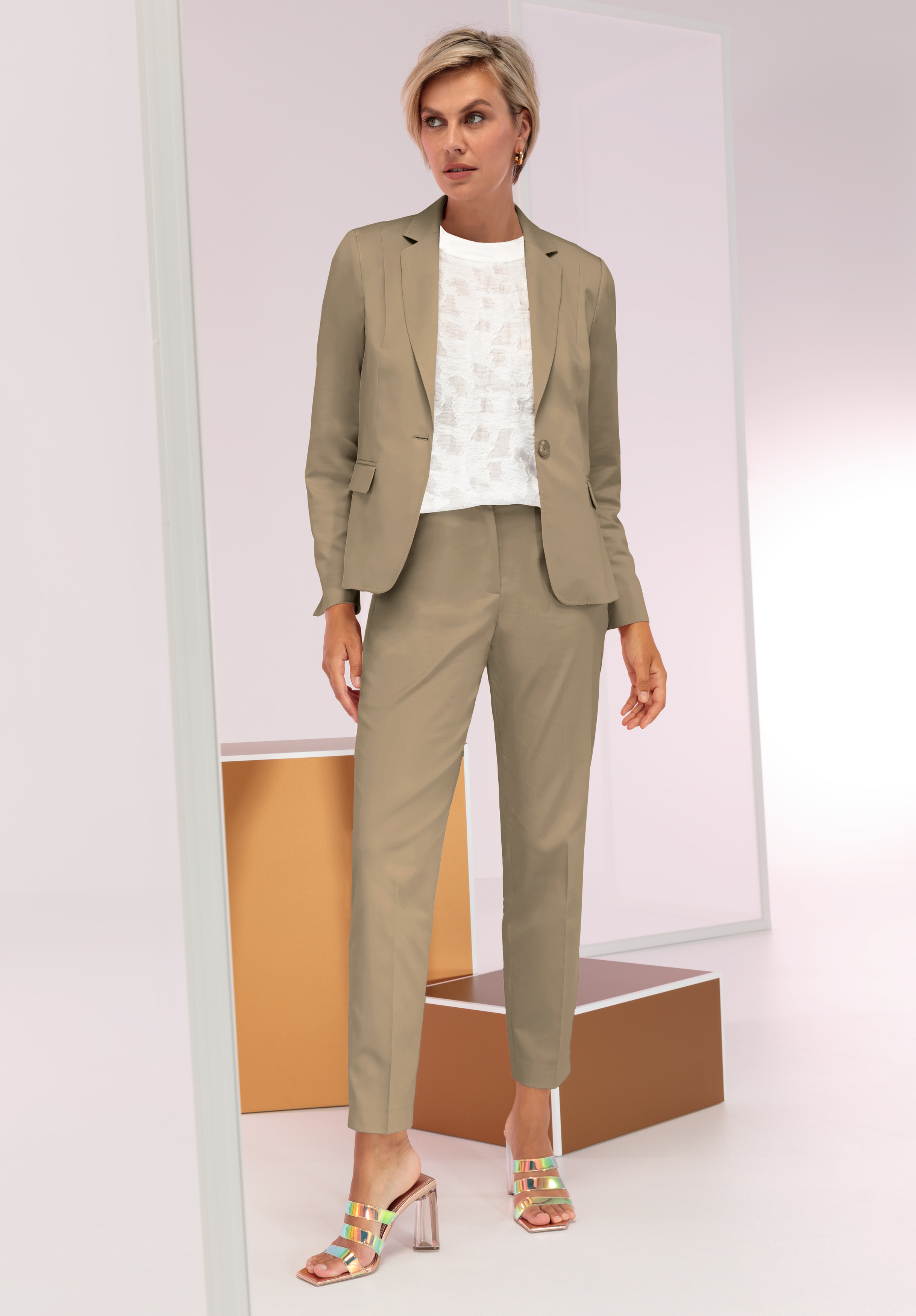 Eine Frau steht in einer minimalistischen, modernen Umgebung. Sie trägt einen hellbraunen Hosenanzug, ein weiß gemustertes Oberteil und schillernde Sandalen mit Absatz.