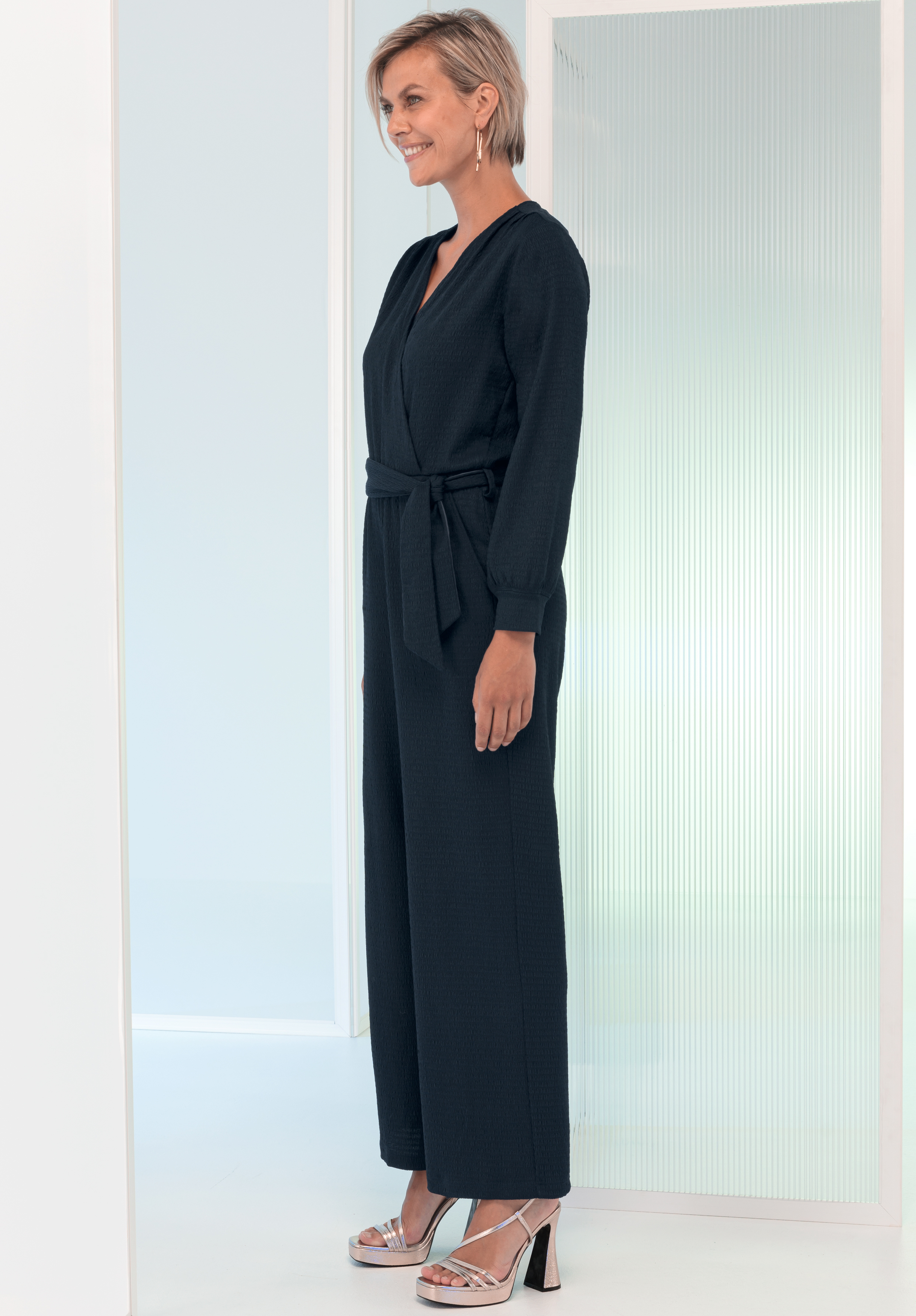 Eine Frau mit kurzen blonden Haaren trägt einen langärmeligen Overall mit dunklem Gürtel und metallische Sandalen mit hohen Absätzen. Sie steht seitlich und lächelt in einem modernen, minimalistischen Raum mit Milchglasplatten.