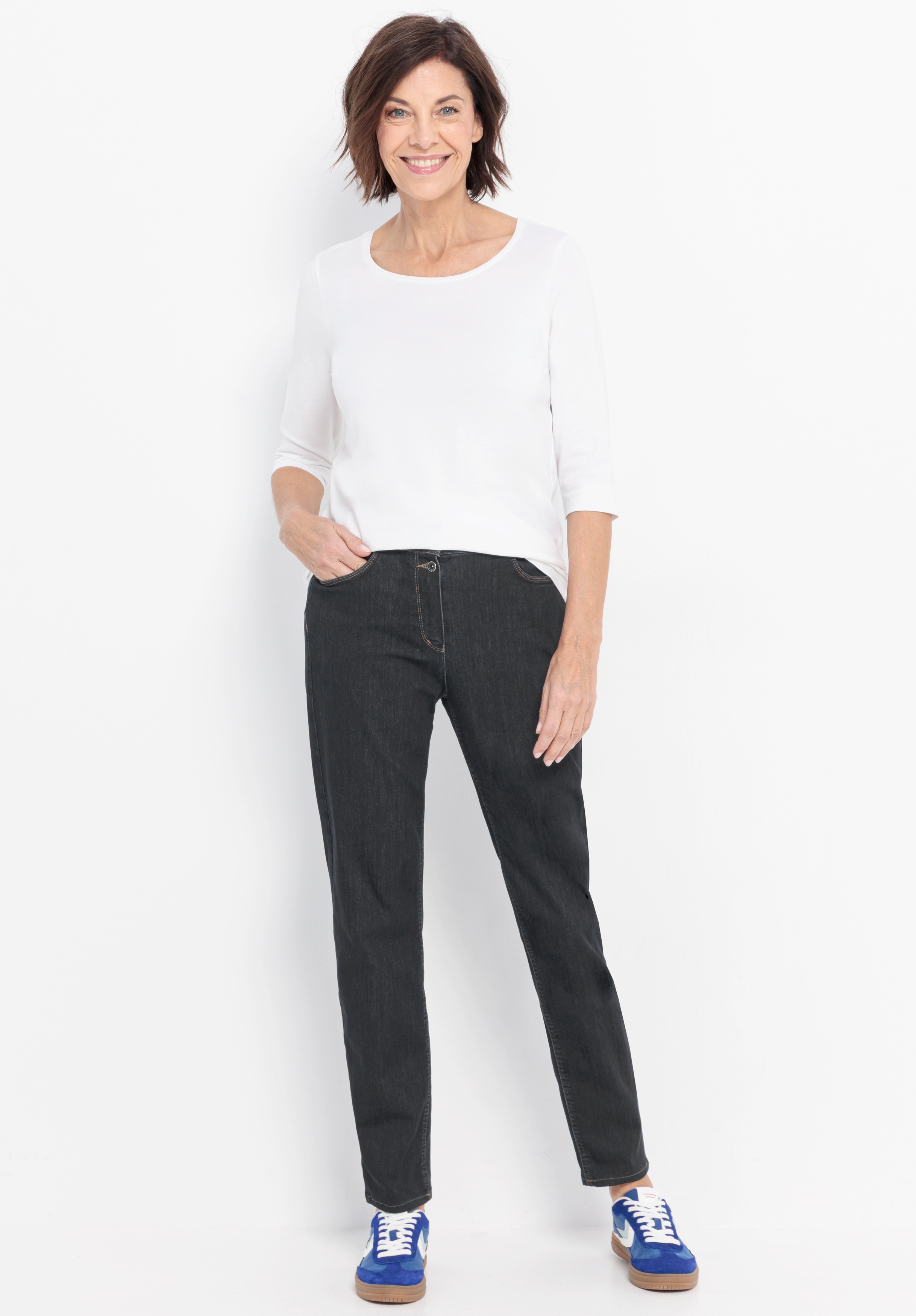 Eine Frau mit kurzen braunen Haaren steht vor einem weißen Hintergrund und lächelt. Sie trägt ein weißes Oberteil mit Dreiviertelärmeln, eine dunkle Jeans und blaue Turnschuhe mit weißen Schnürsenkeln. Eine Hand steckt in ihrer Hosentasche.