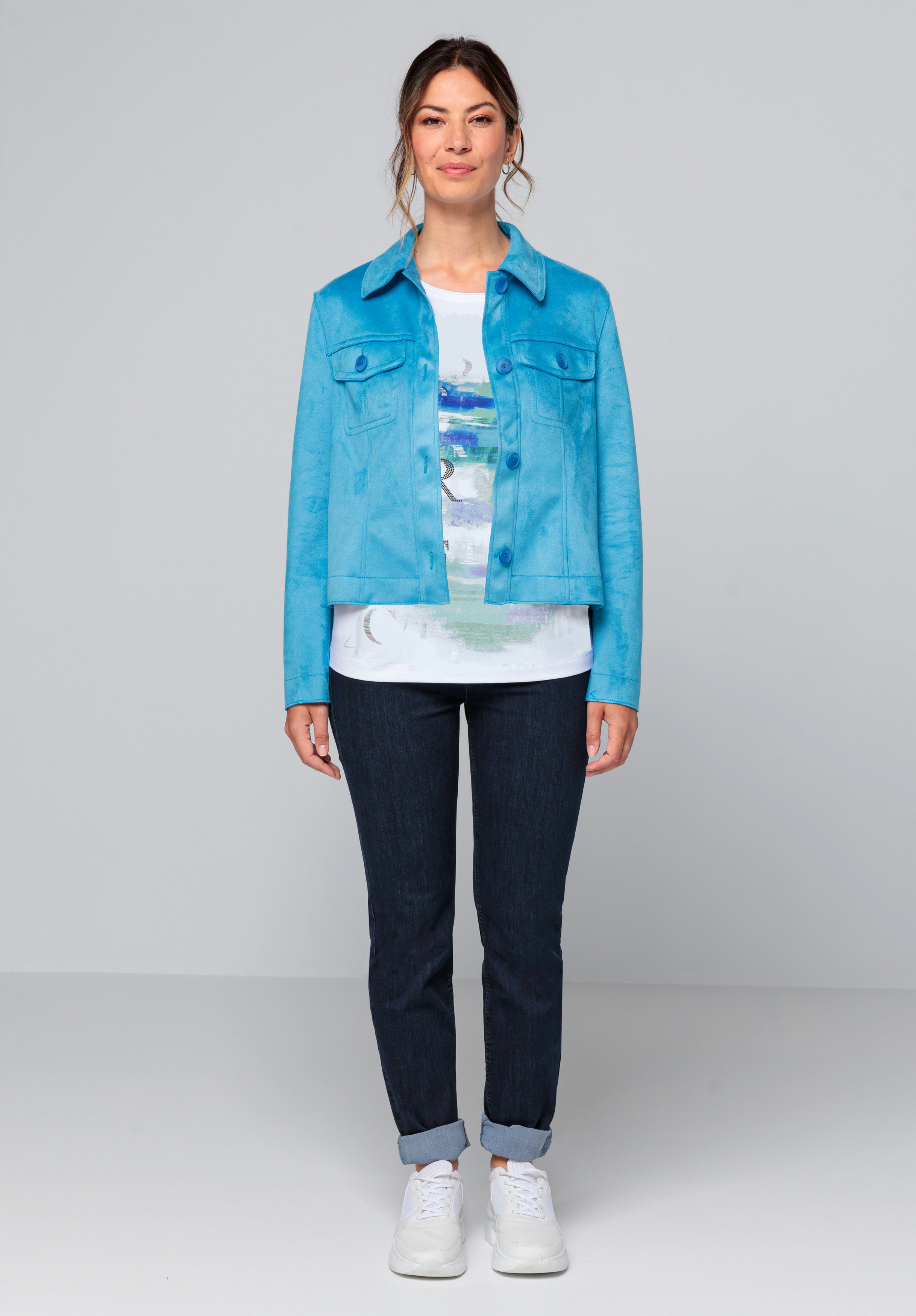 Eine Frau steht mit dem Gesicht nach vorne und trägt eine hellblaue Jacke über einem weißen bedruckten Hemd, dunkle, hochgekrempelte Jeans und weiße Turnschuhe vor einem schlichten hellgrauen Hintergrund.