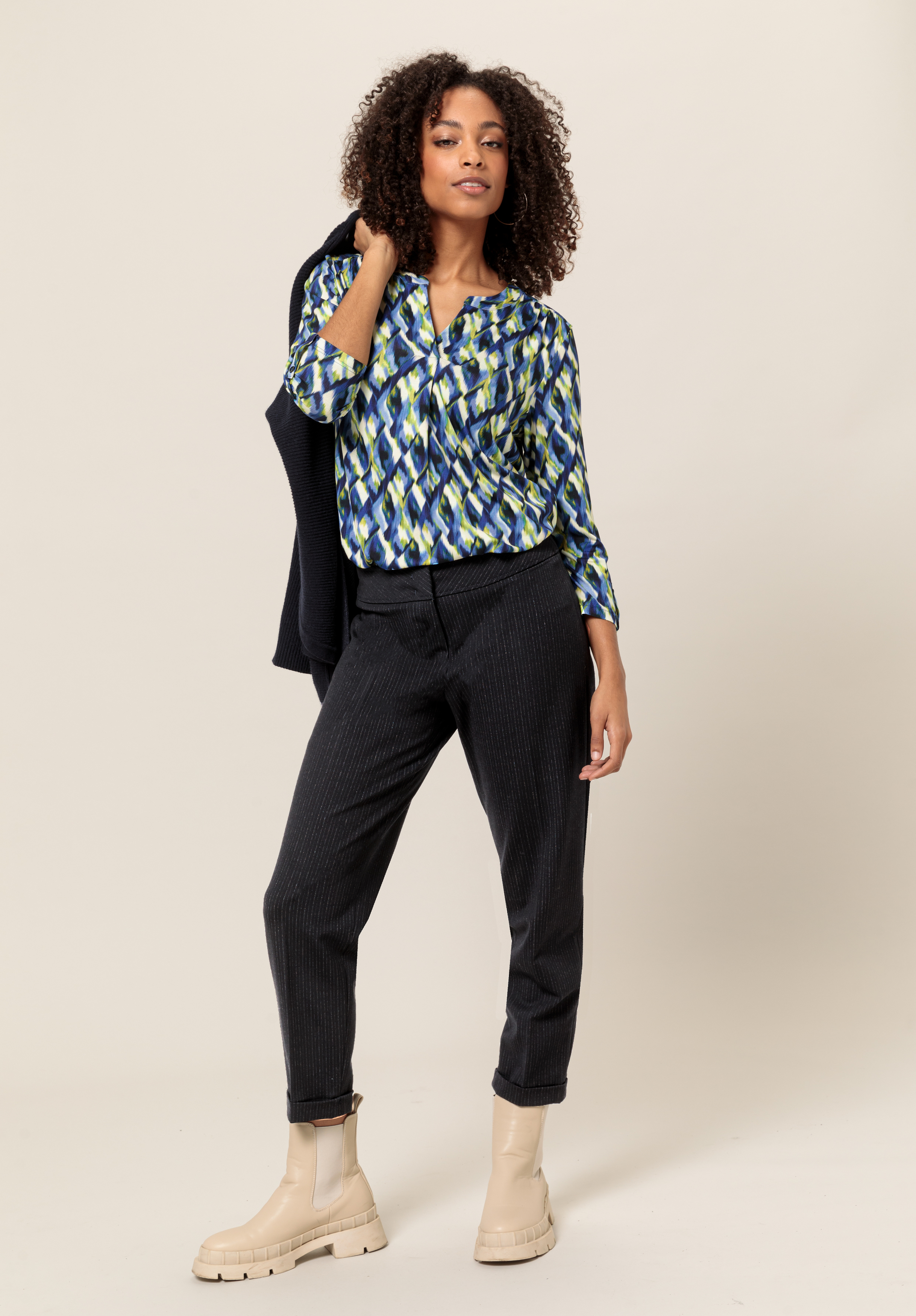 Eine Frau mit lockigem Haar posiert selbstbewusst. Sie trägt eine bunte Bluse mit geometrischem Muster, eine dunkle Hose, hellbeige Stiefel und hält eine dunkle Jacke über der Schulter vor einem neutralen Hintergrund.