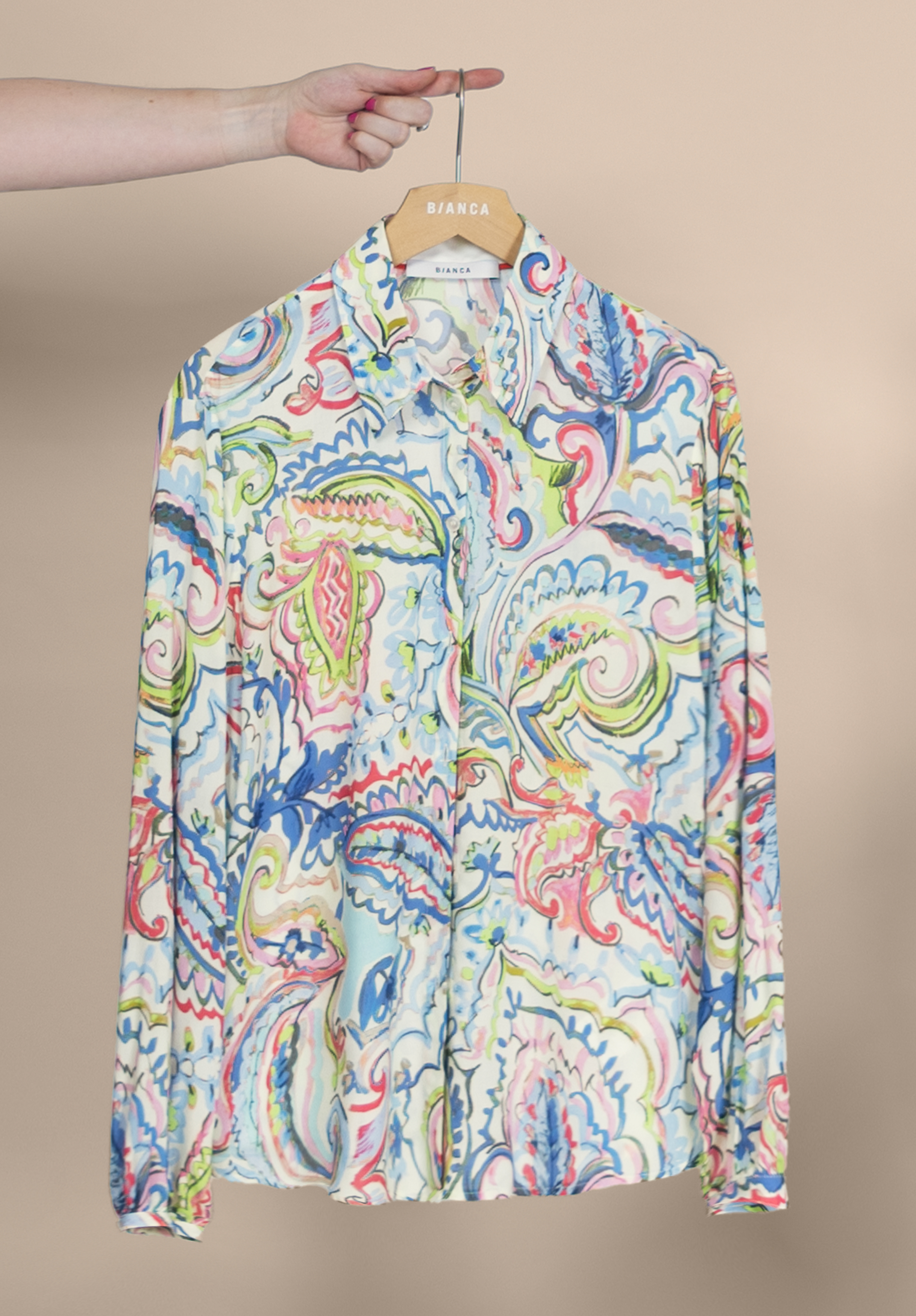 Eine Hand hält einen Kleiderbügel mit einem bunten, langärmeligen Hemd mit Knöpfen, das ein lebhaftes Paisley-Muster in Blau-, Rot-, Grün- und Gelbtönen vor einem neutralen Hintergrund aufweist.