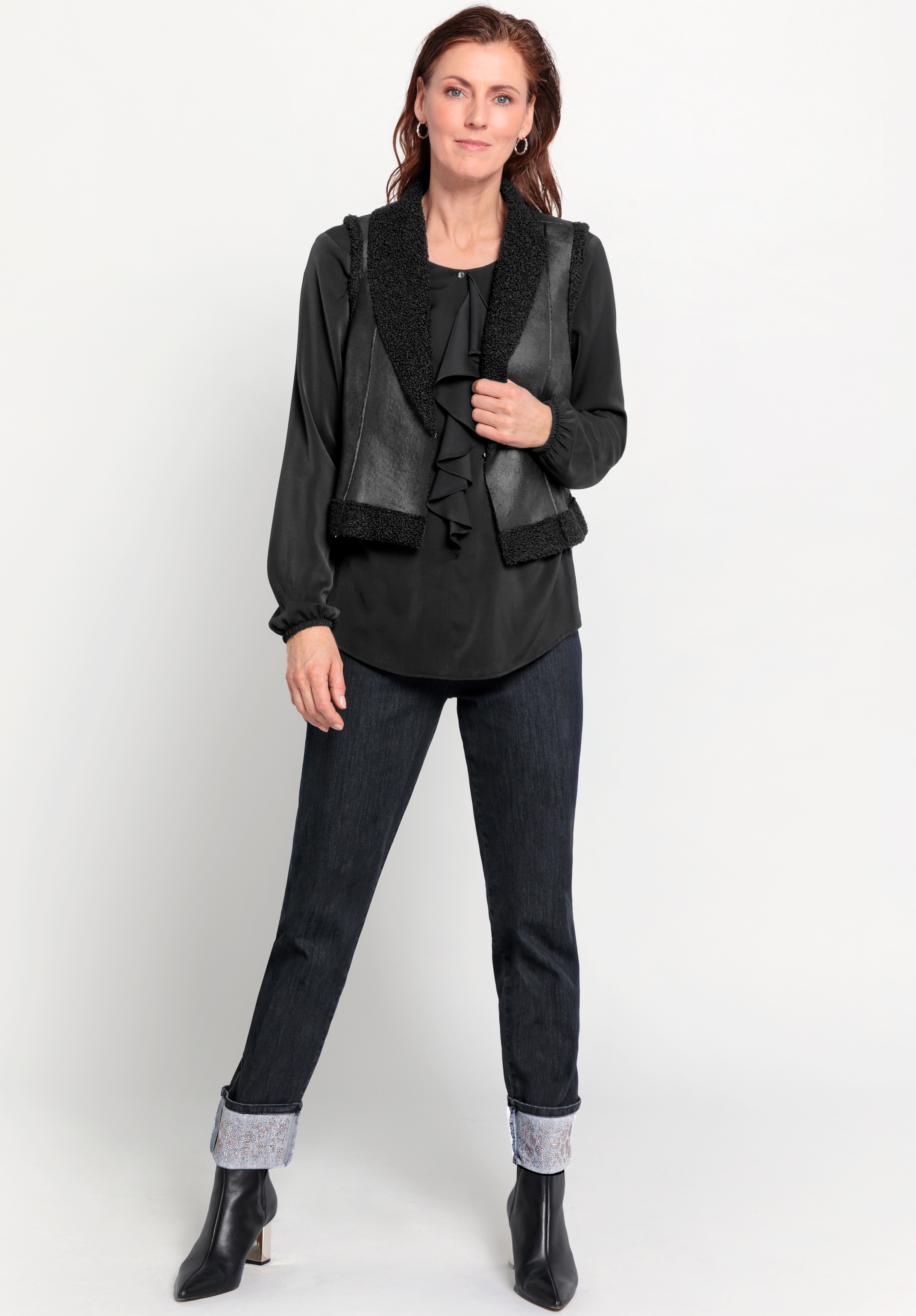 Vor einem weißen Hintergrund steht eine Frau, die eine schwarze Bluse mit Rüschen, eine schwarze, strukturierte Weste, dunkle, hochgekrempelte Jeans und schwarze Stiefeletten trägt. Sie hat langes rotbraunes Haar und eine entspannte, selbstbewusste Haltun