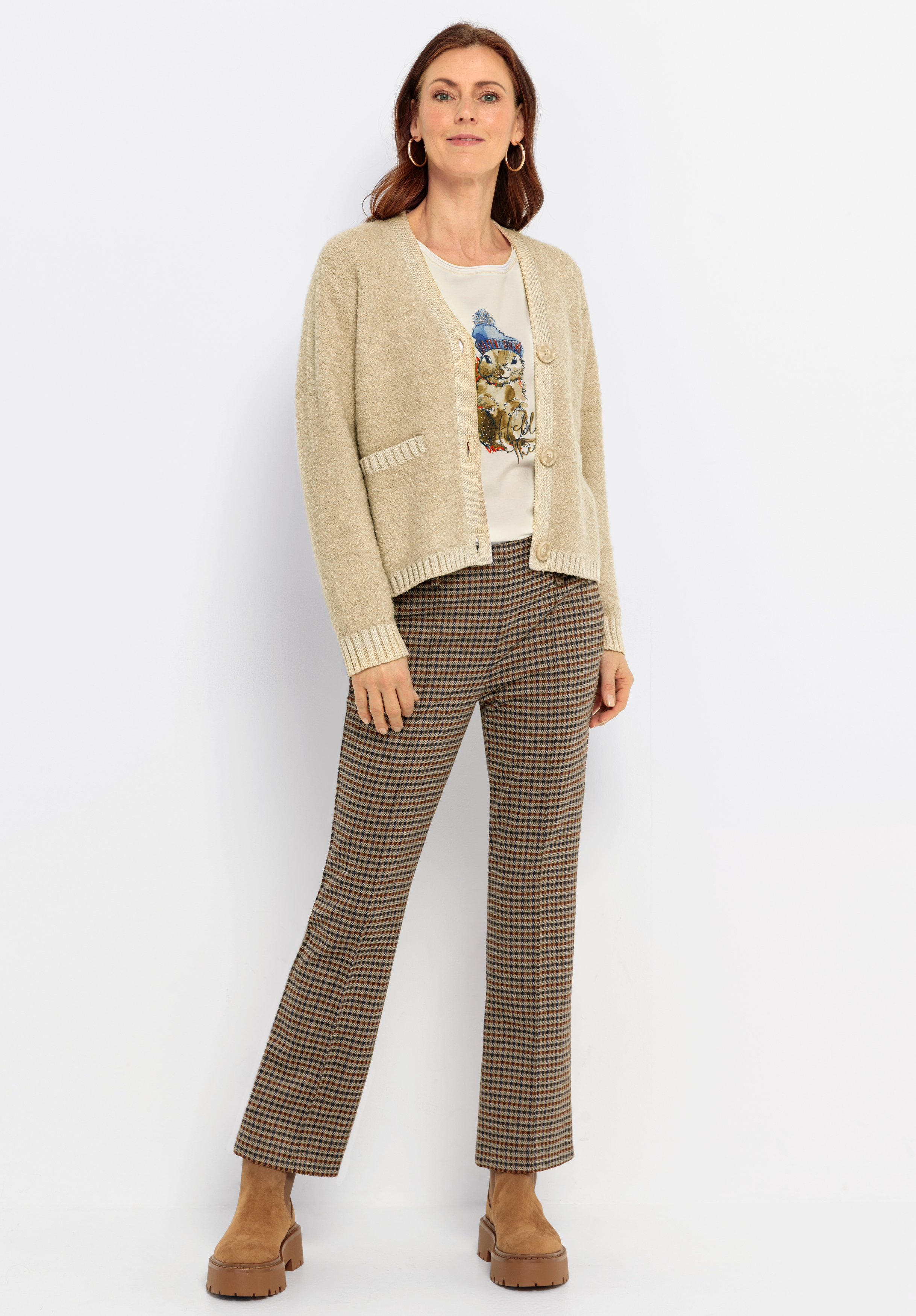 Eine Frau steht vor einem weißen Hintergrund und trägt eine beigefarbene Strickjacke, ein bedrucktes weißes T-Shirt, eine braun karierte Hose und hellbraune Plateauschuhe. Sie hat mittellanges Haar und lächelt sanft in die Kamera.