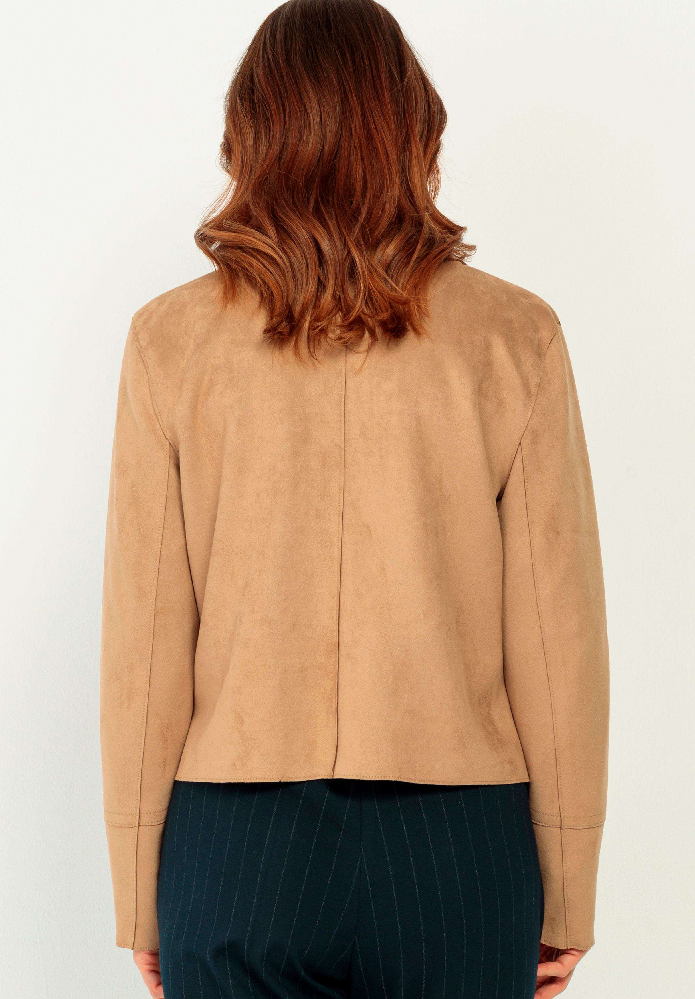 Eine Person mit gewelltem, schulterlangem braunem Haar wird von hinten gezeigt. Sie trägt eine hellbraune Wildlederjacke und eine dunkle Hose mit dünnen, hellen Längsstreifen und steht vor einem schlichten, hellen Hintergrund.