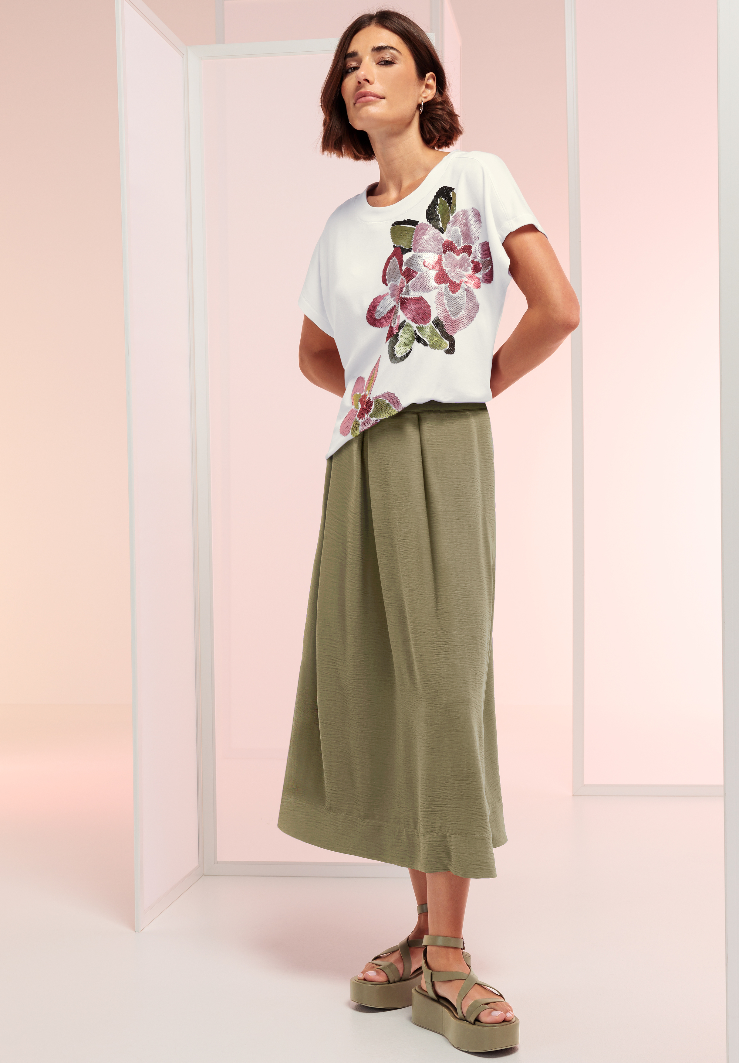 Eine Frau steht in einem weißen Oberteil mit rosa und grünem Blumenmuster, einem olivgrünen Midirock und grünen Plateausandalen in einer minimalistischen, pastellfarbenen Umgebung.