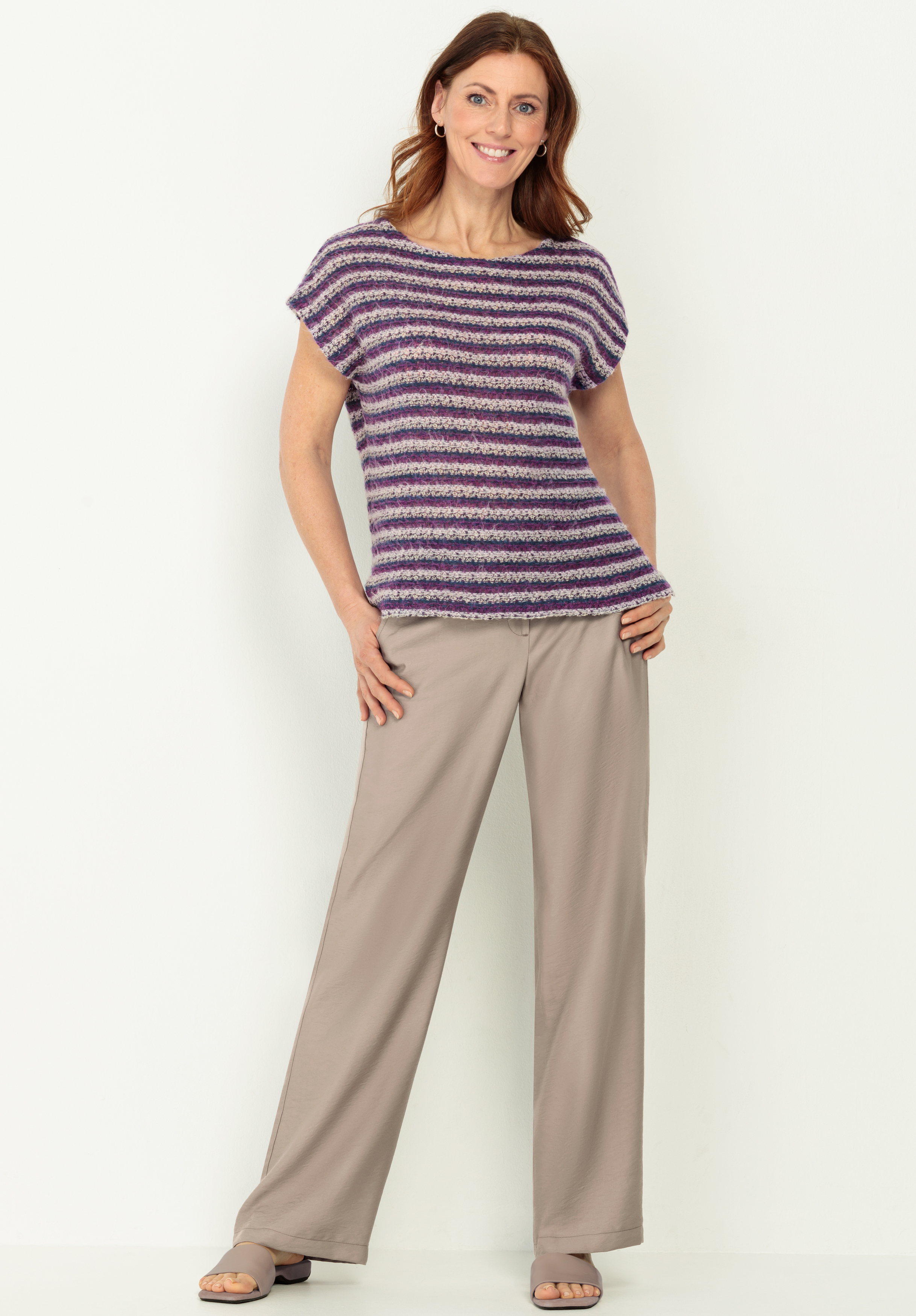 Eine Frau lächelt, während sie ein kurzärmeliges, gestreiftes Strickoberteil in Lila-Tönen, eine weite beigefarbene Hose und Sandalen trägt. Sie steht vor einem schlichten hellen Hintergrund und hat eine Hand in die Hüfte gestemmt.