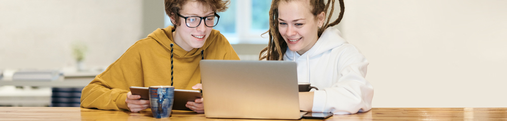 Zwei lächelnde junge Menschen sitzen an einem Holztisch und schauen auf einen Laptop. Einer hält ein Tablet in der Hand, trägt eine Brille und einen gelben Kapuzenpullover, der andere einen weißen. Vor ihnen steht eine blaue Tasse.