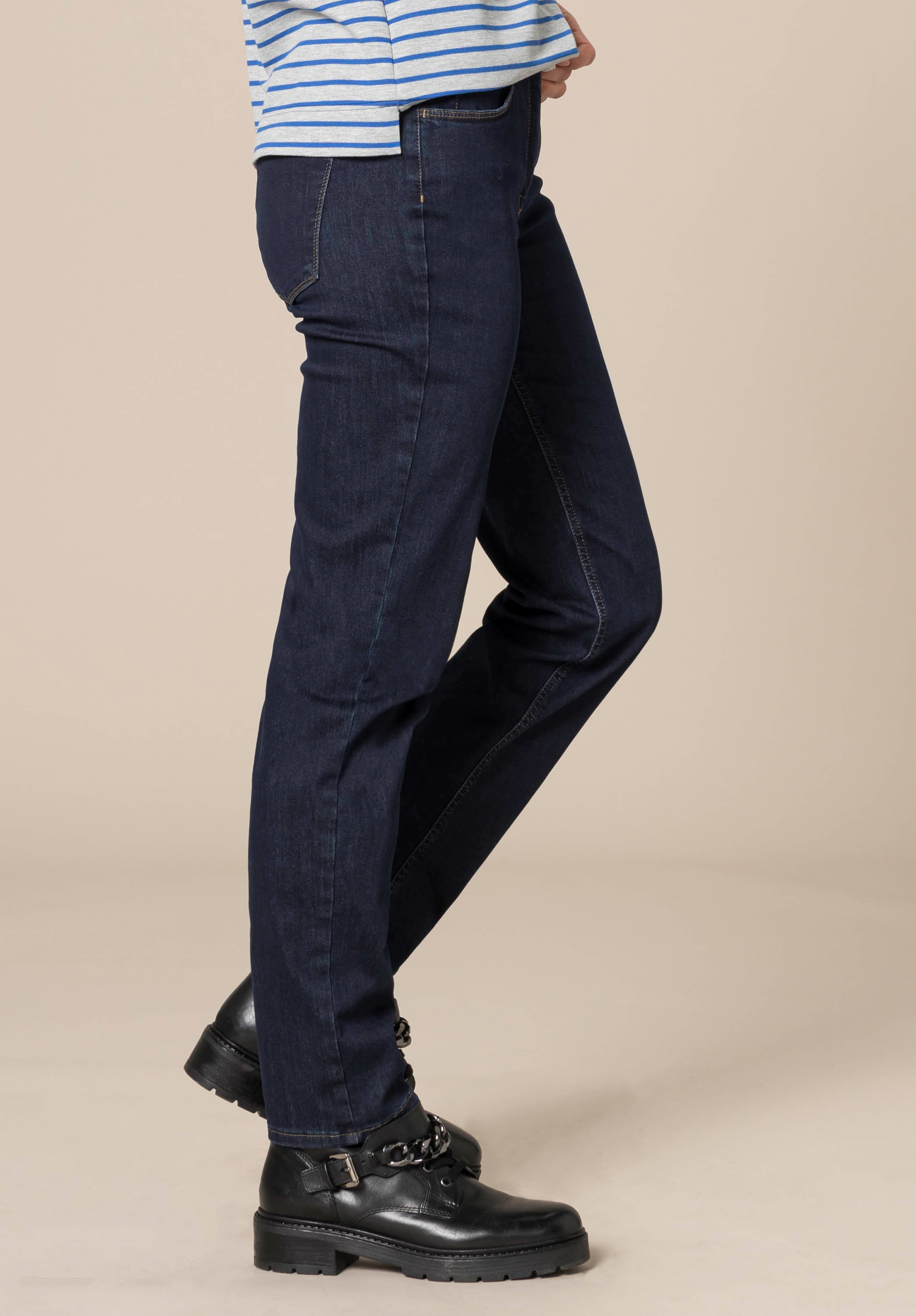 Person, die dunkelblaue Jeans mit geradem Bein, ein blau-weiß gestreiftes Oberteil und schwarze Schnürschuhe trägt und vor einem schlichten beigen Hintergrund steht. Der Oberkörper ist größtenteils außerhalb des Bildausschnitts.