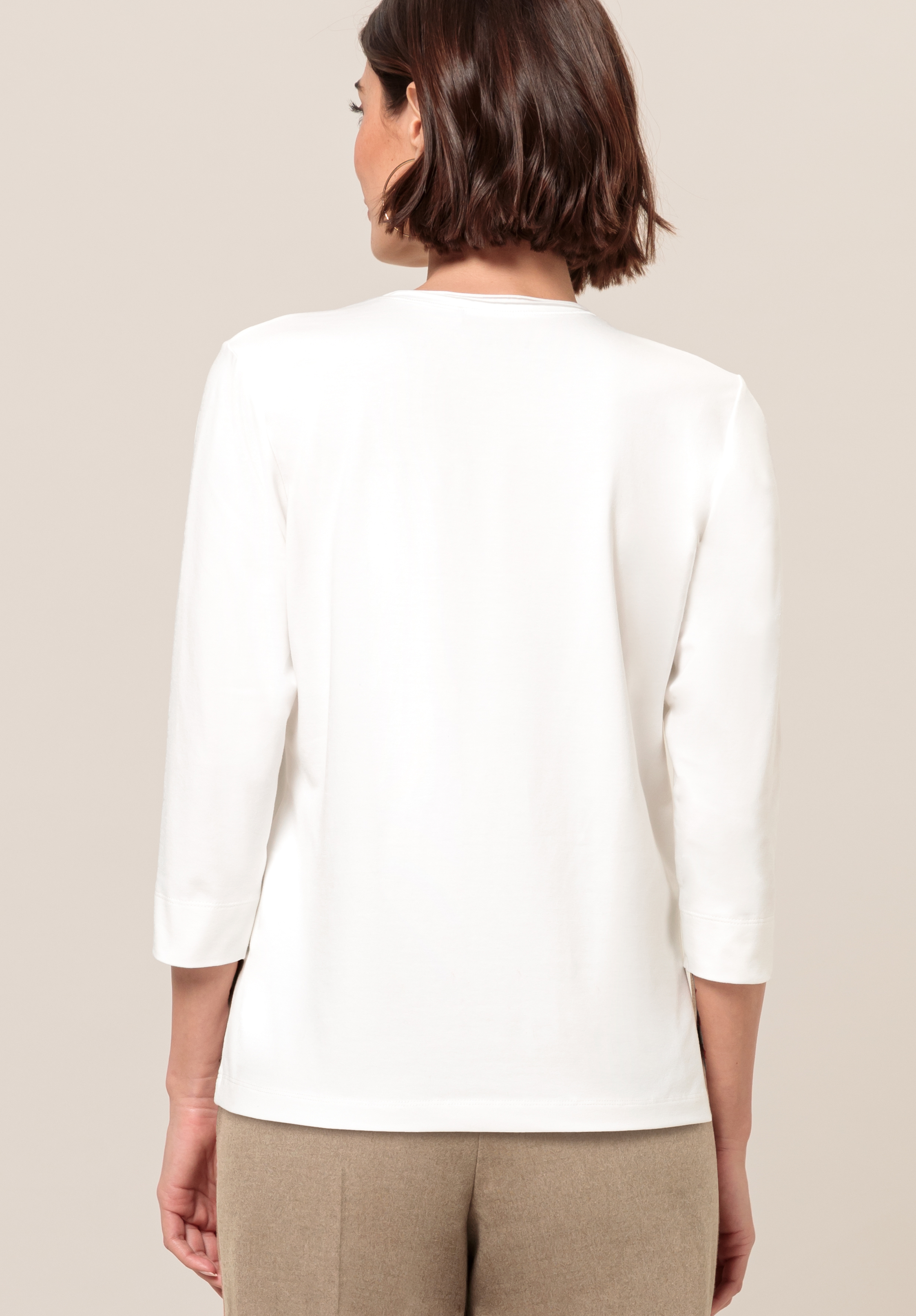 Von hinten ist eine Frau mit kurzen braunen Haaren zu sehen, die vor einem schlichten, hellen Hintergrund ein weißes Oberteil mit Dreiviertelärmeln und eine beige Hose trägt.
