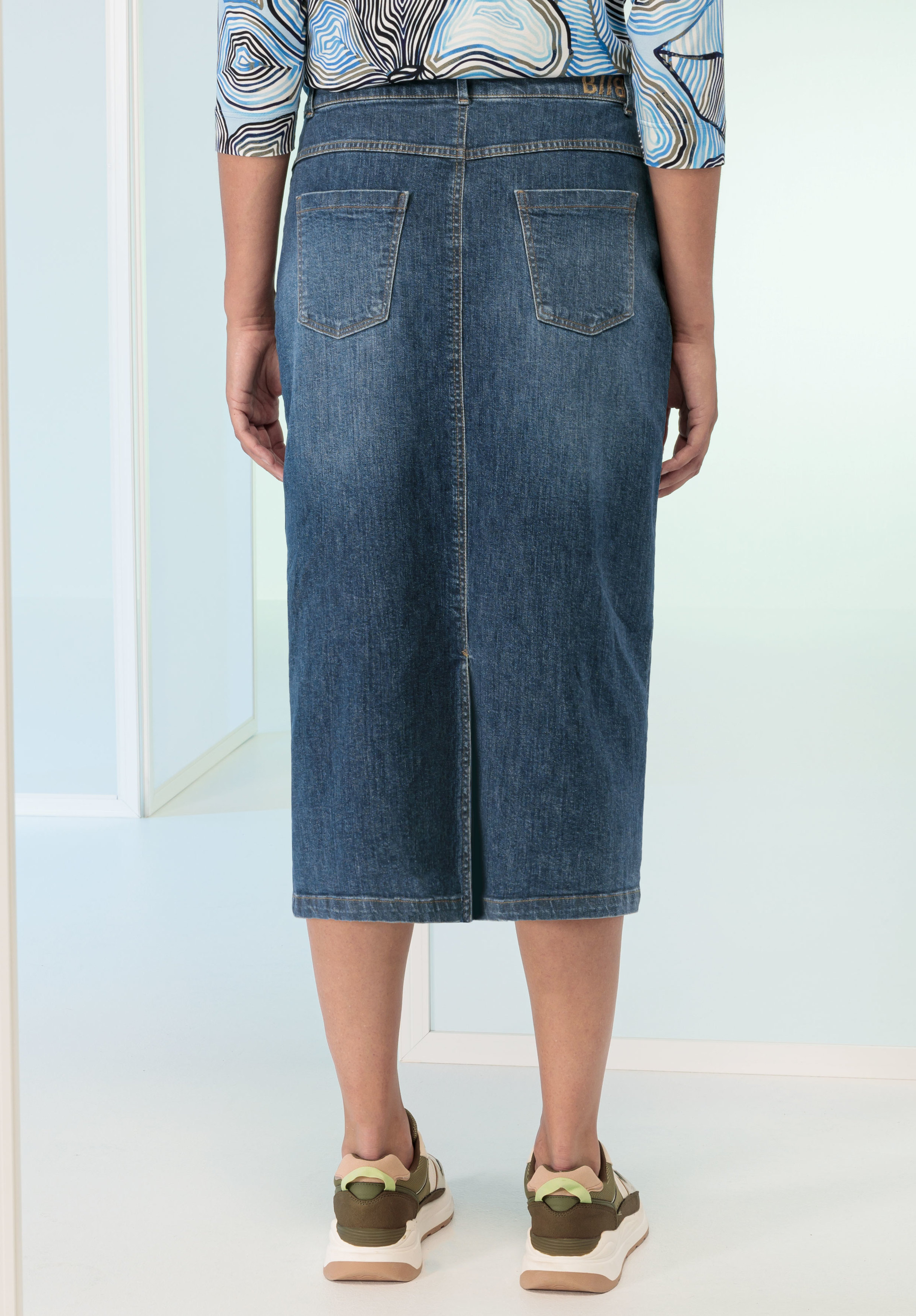 Eine Person in einem knielangen Jeansrock und Sneakers steht mit dem Rücken zur Kamera. Der Rock hat zwei Gesäßtaschen, die Person trägt ein gemustertes Langarmshirt.