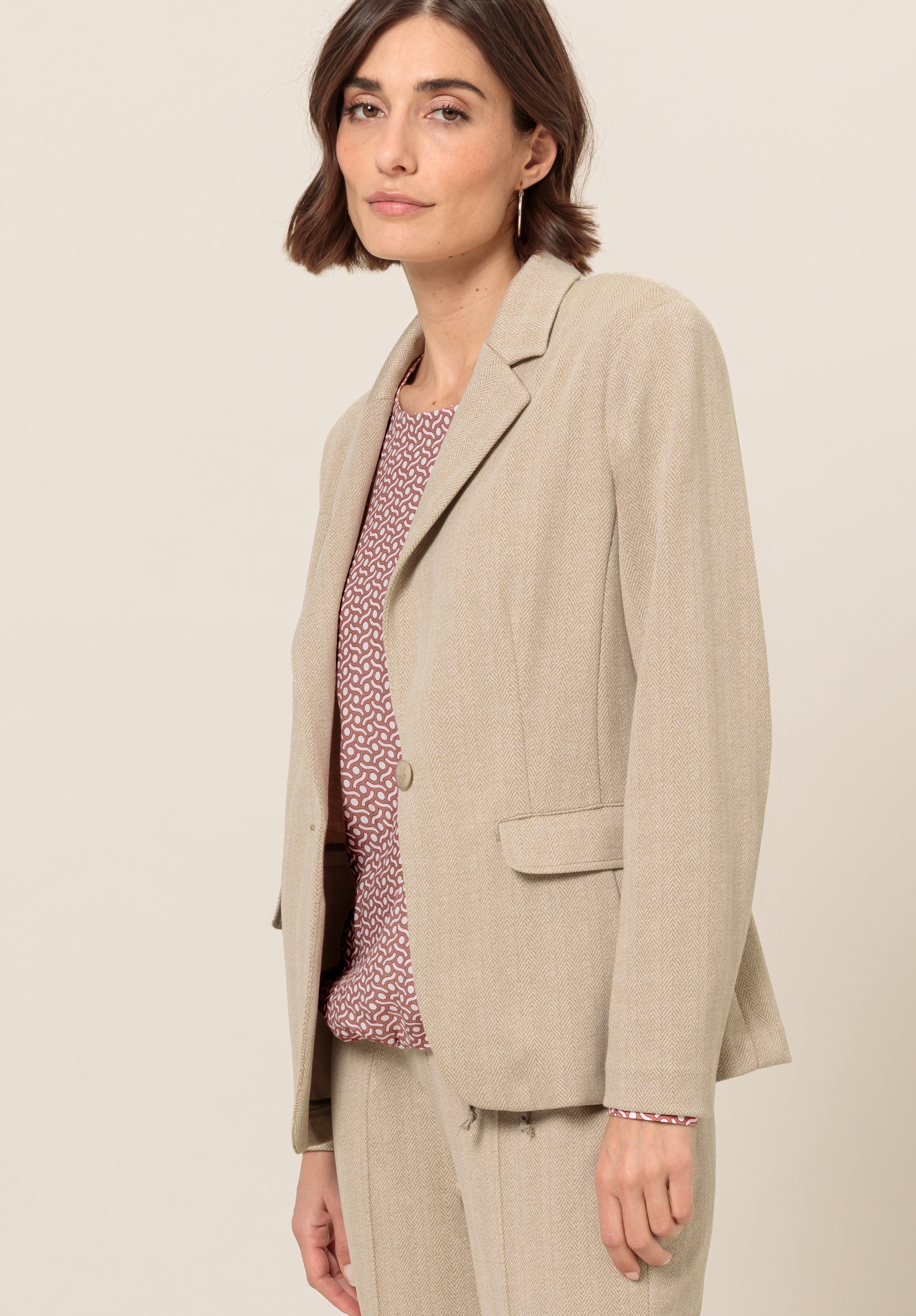 Eine Frau mit kurzen braunen Haaren trägt einen beigen Blazer und eine passende Hose über einer gemusterten rosa Bluse, steht vor einem schlichten hellen Hintergrund und blickt mit neutralem Gesichtsausdruck in die Kamera.