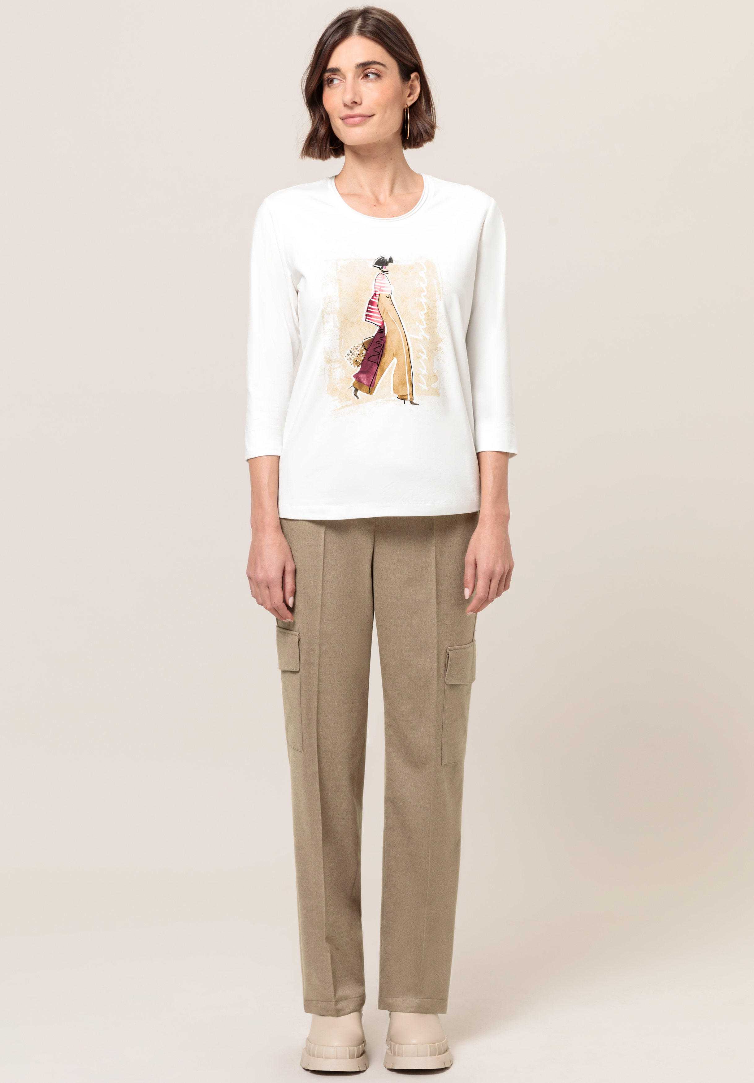 Eine Frau steht mit dem Gesicht nach vorne. Sie trägt ein weißes Langarmshirt mit künstlerischem Aufdruck und beige Cargohosen. Sie hat kurze braune Haare und trägt helle Schuhe vor einem schlichten hellen Hintergrund.