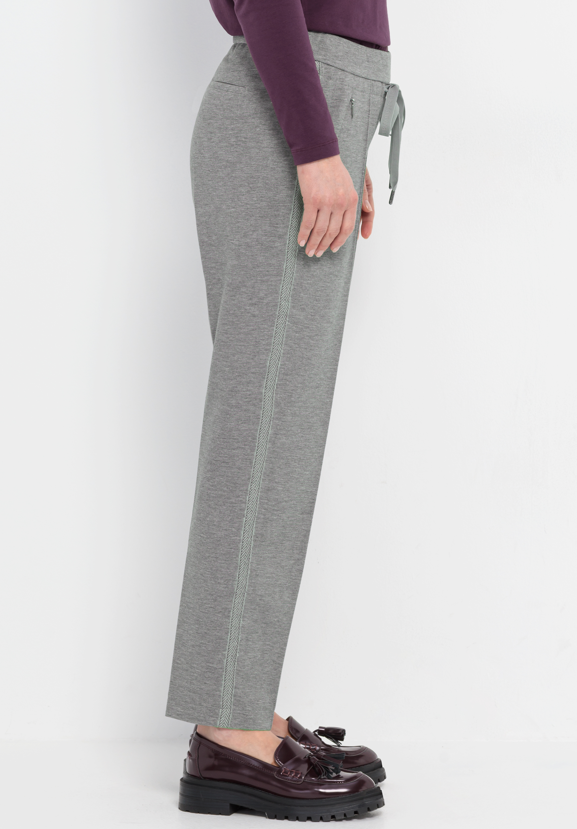Eine Person steht seitlich und trägt eine graue Hose mit geradem Bein und Kordelzug, ein langärmeliges lila Oberteil und kastanienbraune Slipper mit Quasten vor einem schlichten weißen Hintergrund.