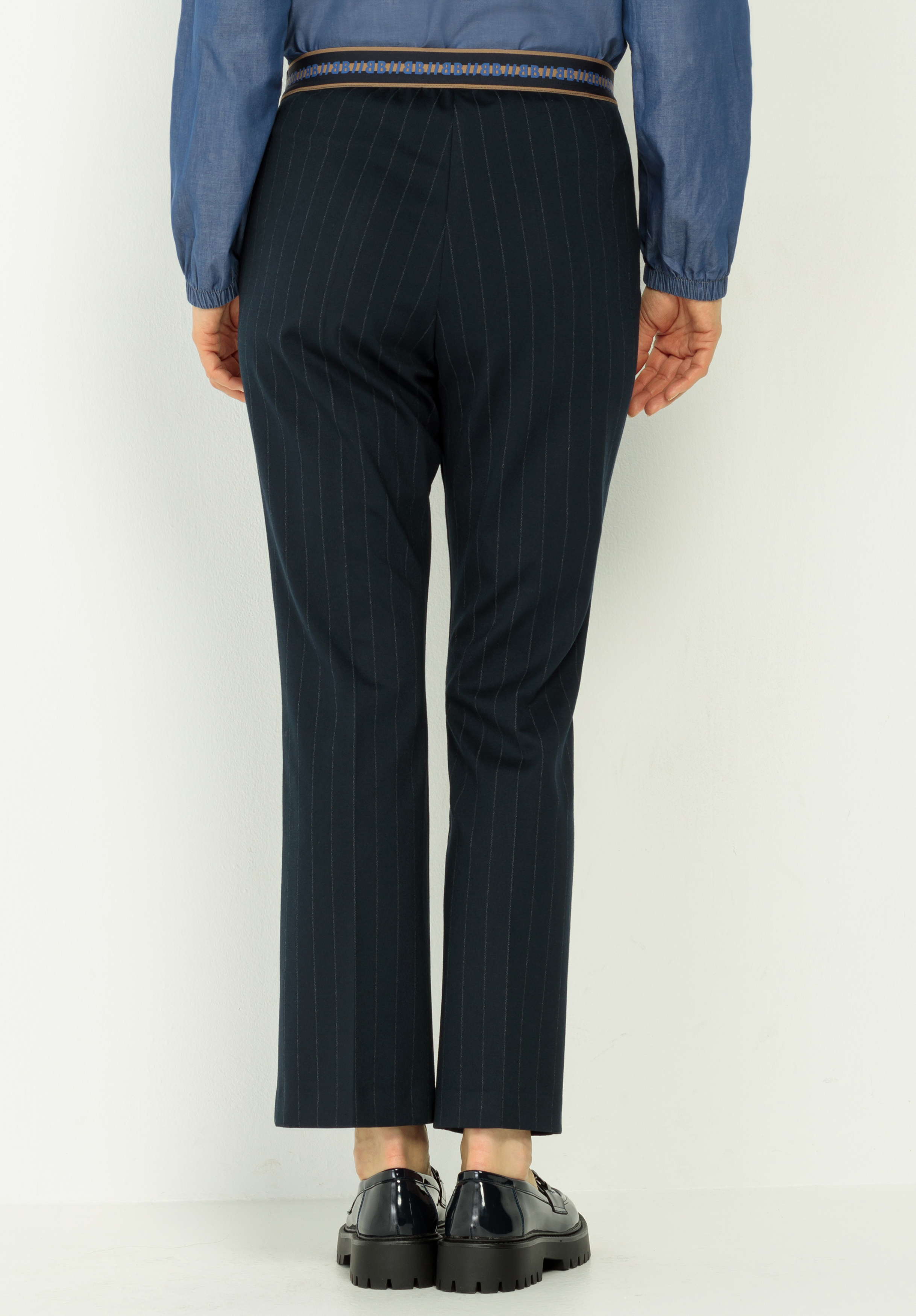 Eine Person, die eine schwarze Nadelstreifenhose, ein blaues langärmeliges Hemd und schwarze Halbschuhe trägt, steht mit dem Rücken zur Kamera vor einem schlichten weißen Hintergrund.