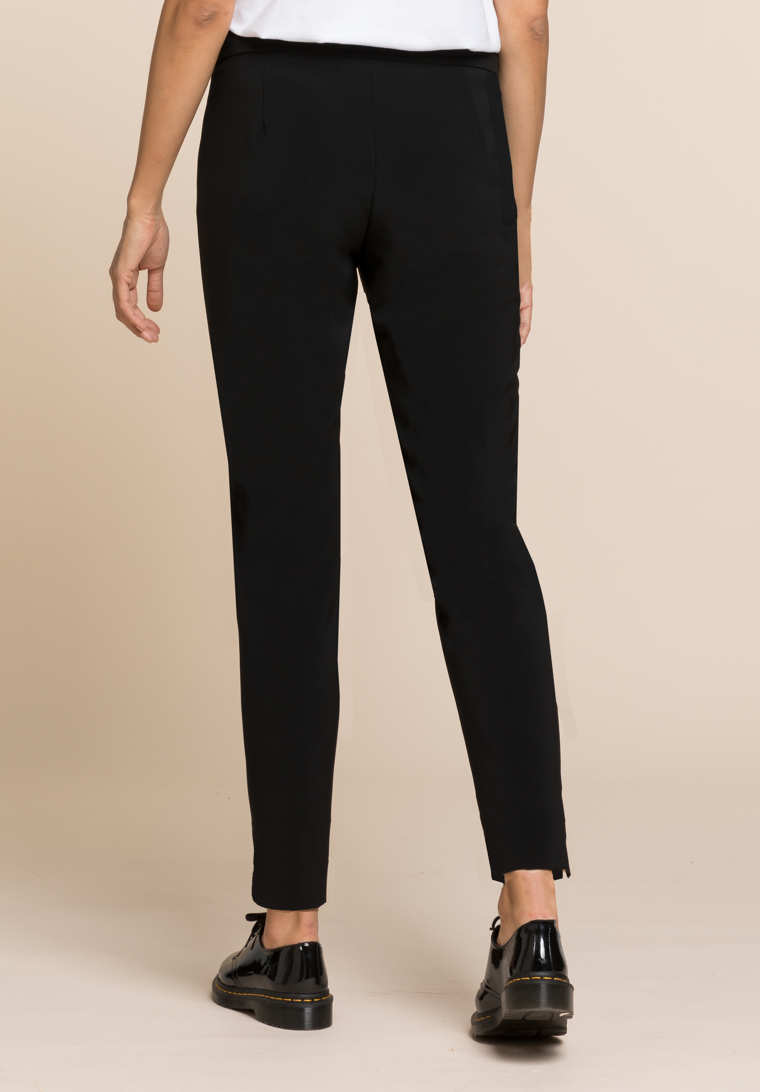 Eine Person steht vor einem beigen Hintergrund; sie ist von der Taille abwärts zu sehen und trägt eine schwarze, eng anliegende Hose sowie schwarze, glänzende Schuhe mit Quasten.
