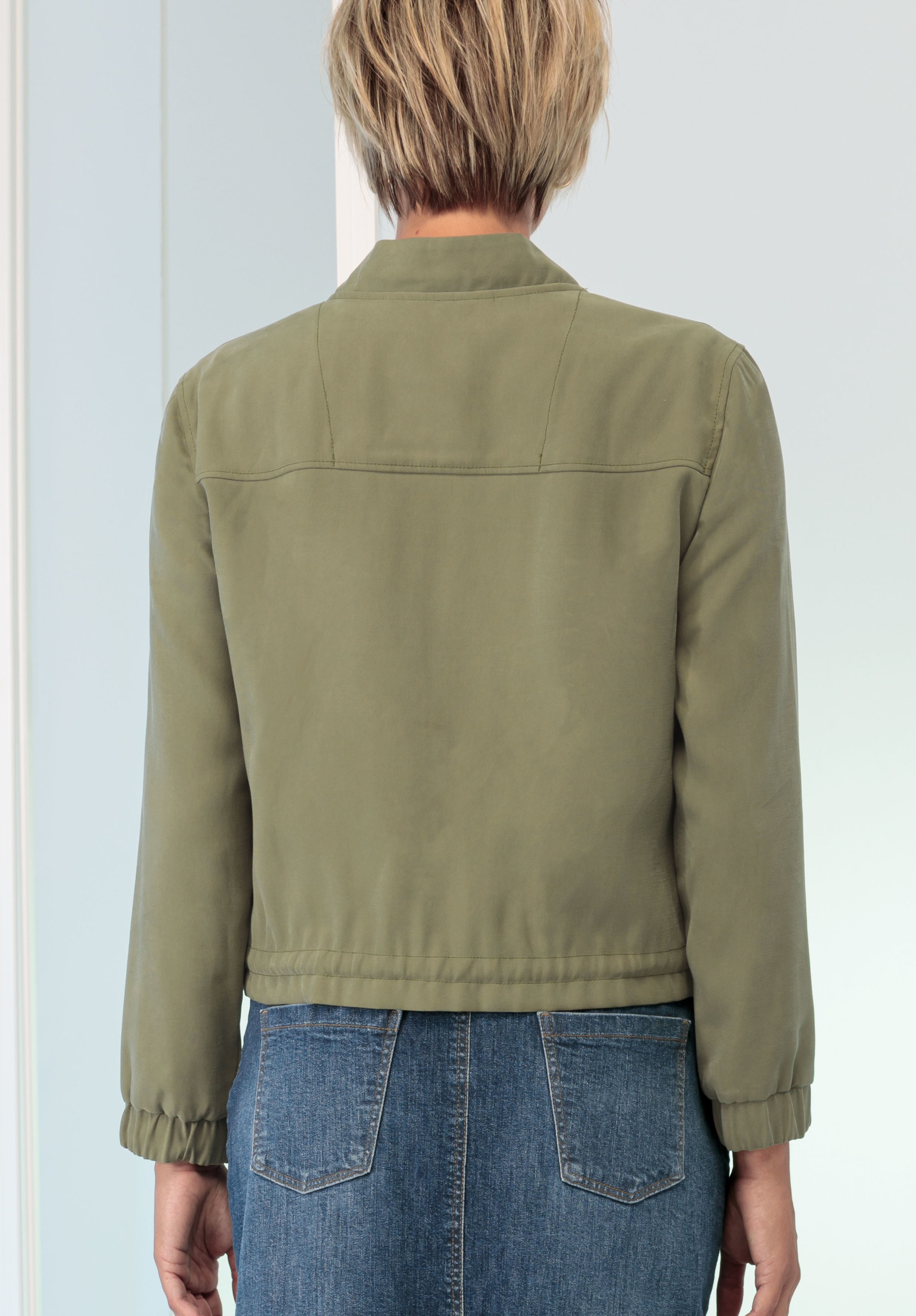 Eine Person mit kurzen, blonden Haaren ist von hinten zu sehen. Sie trägt eine hell olivgrüne Jacke und blaue Jeans. Der Hintergrund ist sanft beleuchtet und neutral.