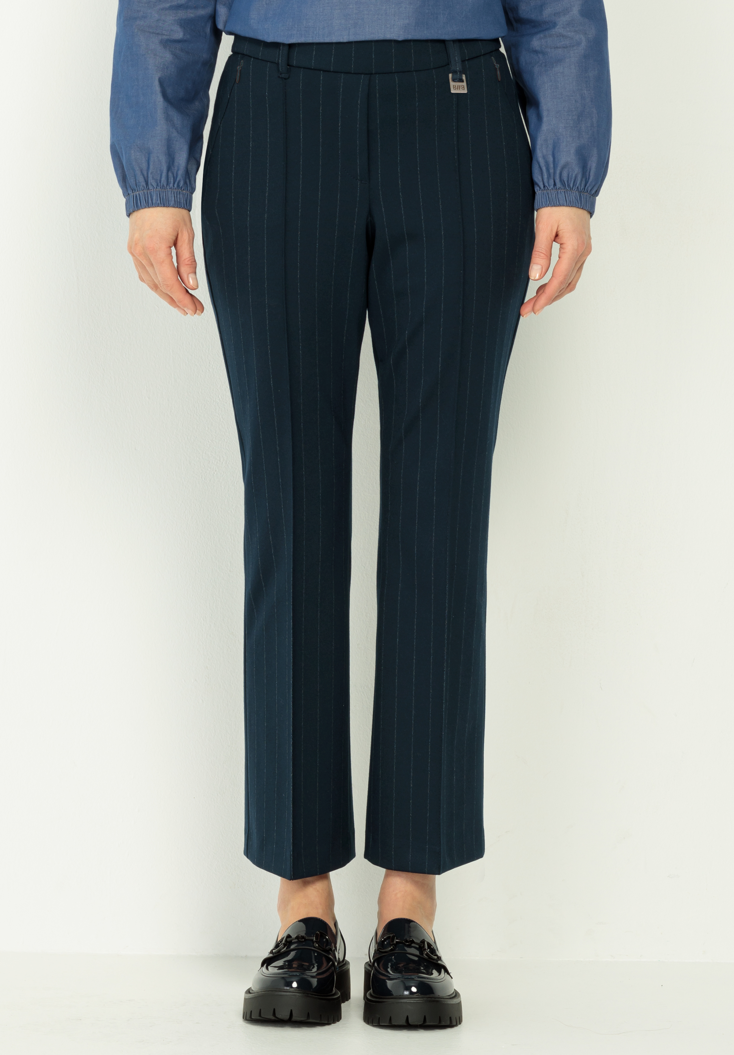 Eine Person, die eine dunkle Nadelstreifenhose, ein blaues langärmeliges Hemd und schwarze glänzende Slipper trägt, steht vor einem schlichten hellen Hintergrund. Das Bild zeigt das Outfit von den Schultern bis zu den Füßen.