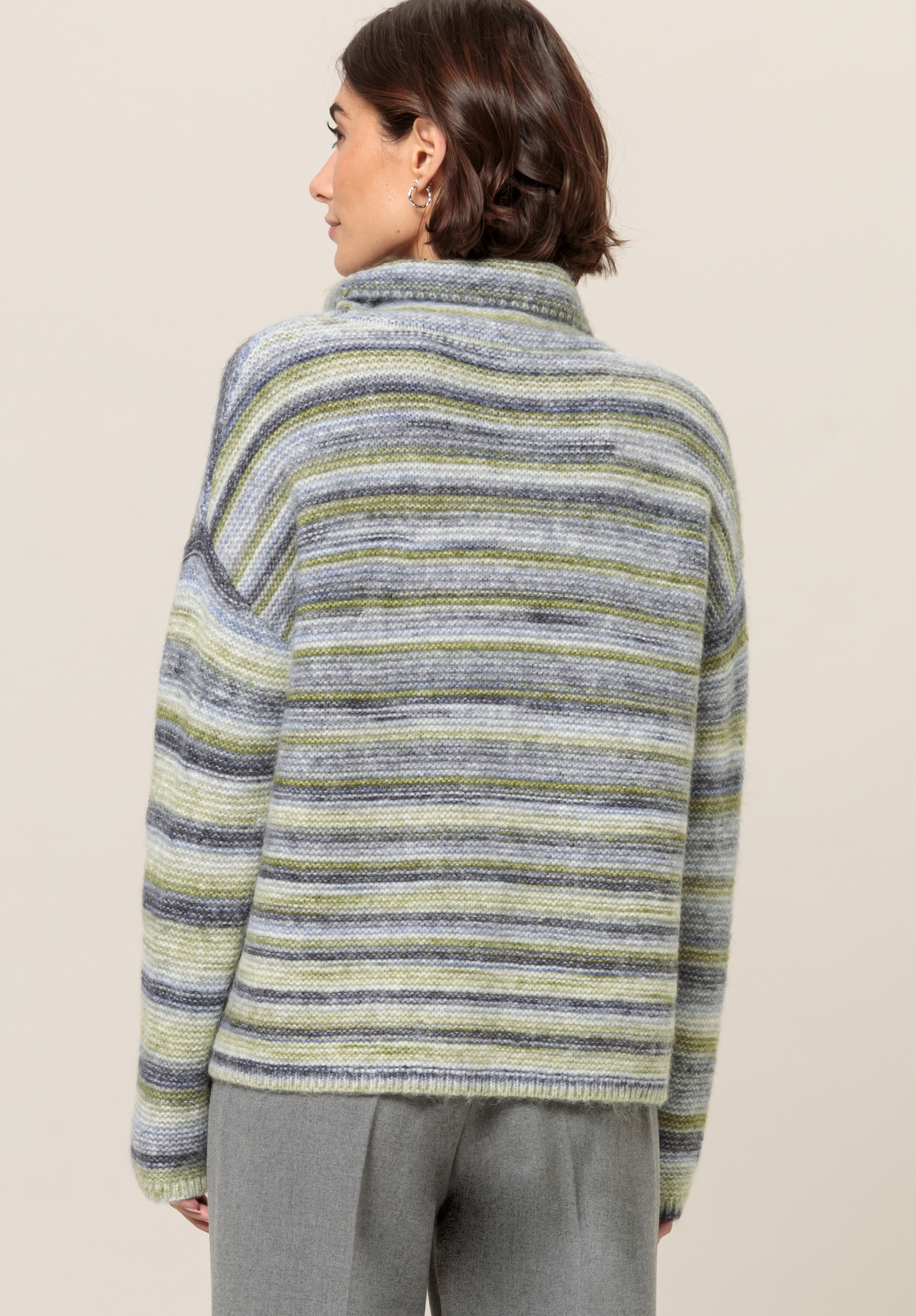 Von hinten ist eine Person mit kurzen braunen Haaren zu sehen, die einen gestreiften Pullover in Grau-, Grün- und Blautönen und eine graue Hose vor einem schlichten beigen Hintergrund trägt.