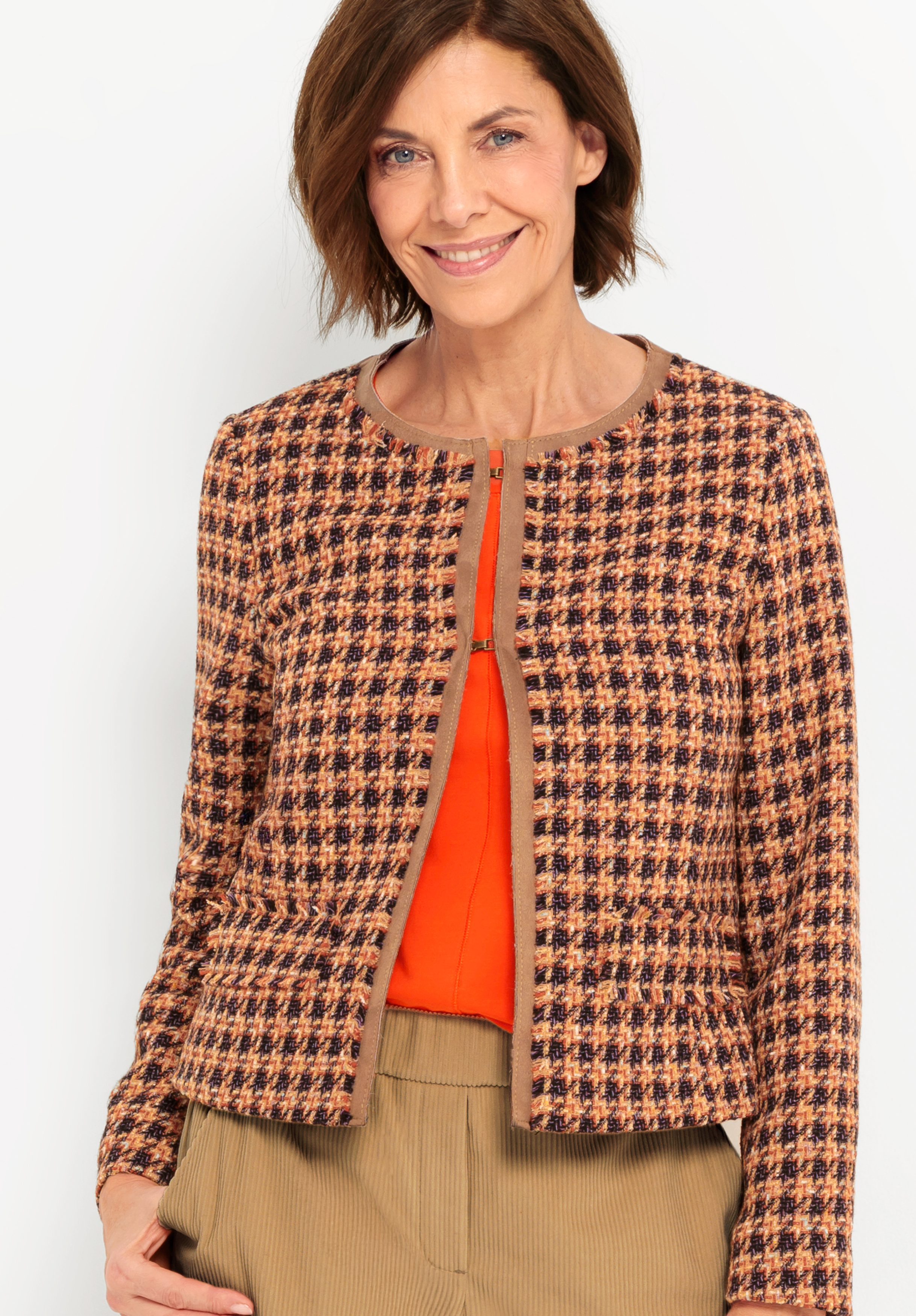 Eine Frau mit kurzen braunen Haaren lächelt, während sie eine karierte Jacke in Braun- und Orangetönen über einem leuchtend orangefarbenen Oberteil trägt, das mit einer hellbraunen Hose kombiniert ist. Sie steht vor einem schlichten, hellen Hintergrund.