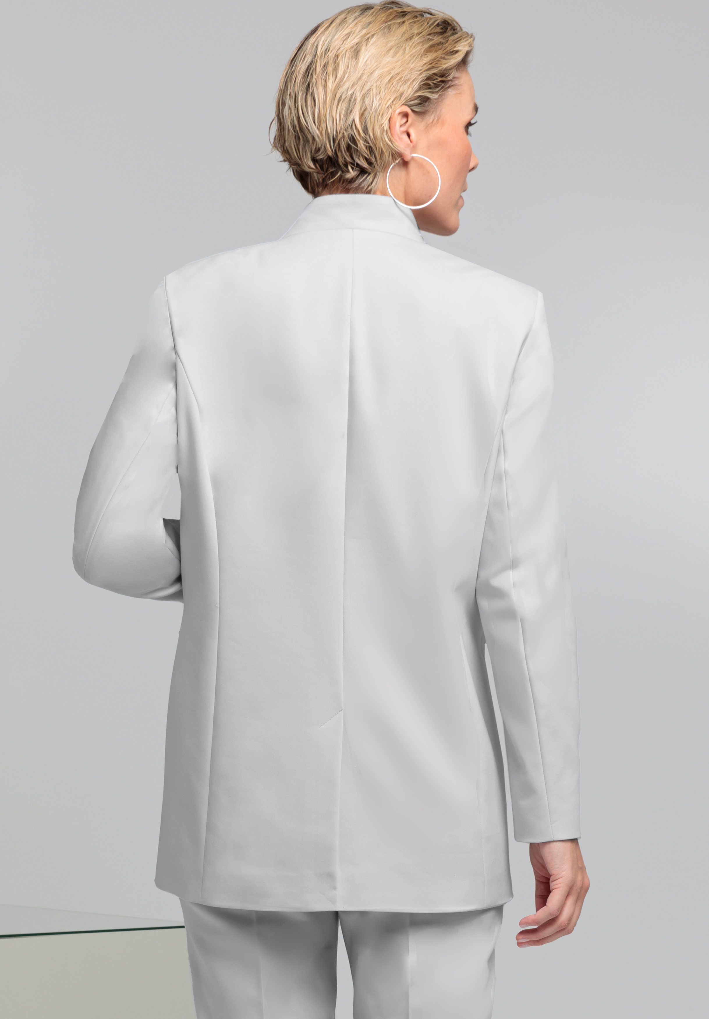Eine Person mit kurzen, blonden Haaren, einem weißen Jackett und der dazu passenden Hose ist von hinten zu sehen. Sie steht vor einem schlichten hellgrauen Hintergrund. Sie trägt einen großen Creolenohrring.
