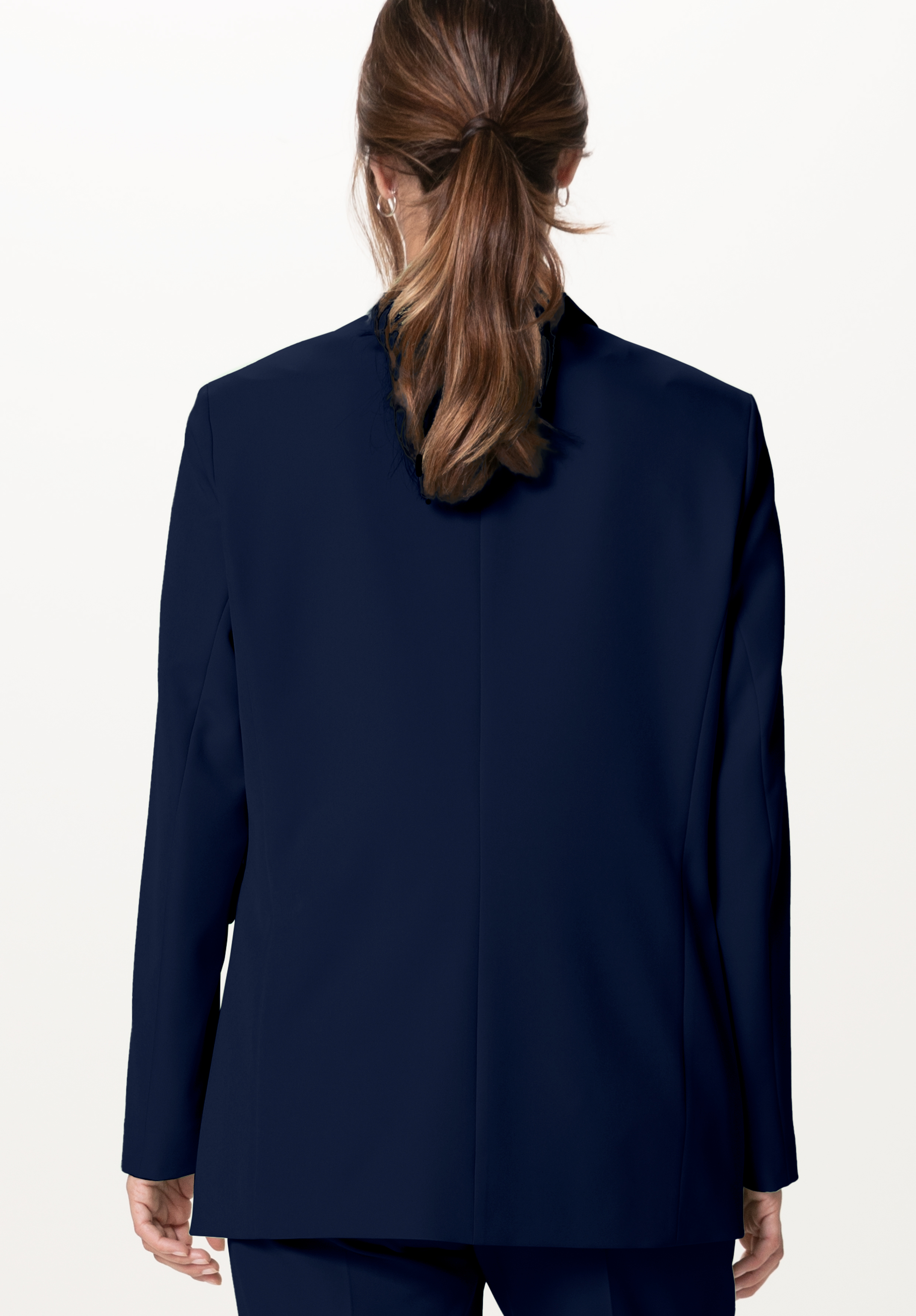 Eine Person mit braunem Haar in einem niedrigen Pferdeschwanz wird von hinten gezeigt. Sie trägt eine dunkle marineblaue Anzugsjacke und eine passende Hose vor einem schlichten hellen Hintergrund.