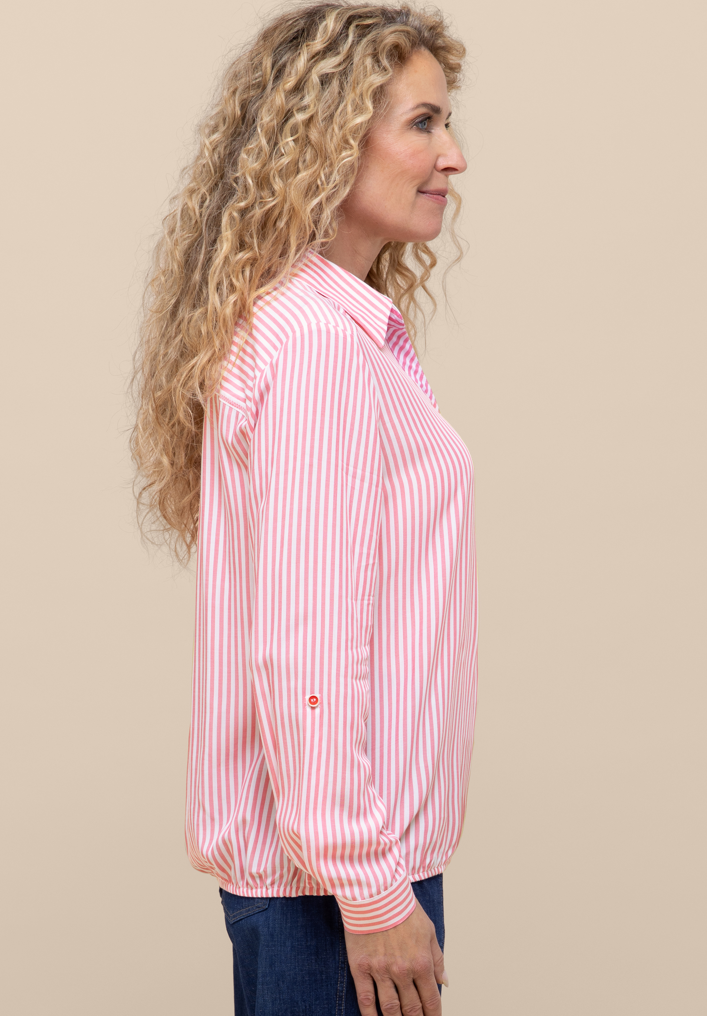 Vor einem schlichten beigen Hintergrund steht eine Frau im Profil mit langen, lockigen blonden Haaren; sie trägt eine rosa-weiß vertikal gestreifte Bluse mit umgeschlagenen Ärmelbündchen und eine blaue Jeans.