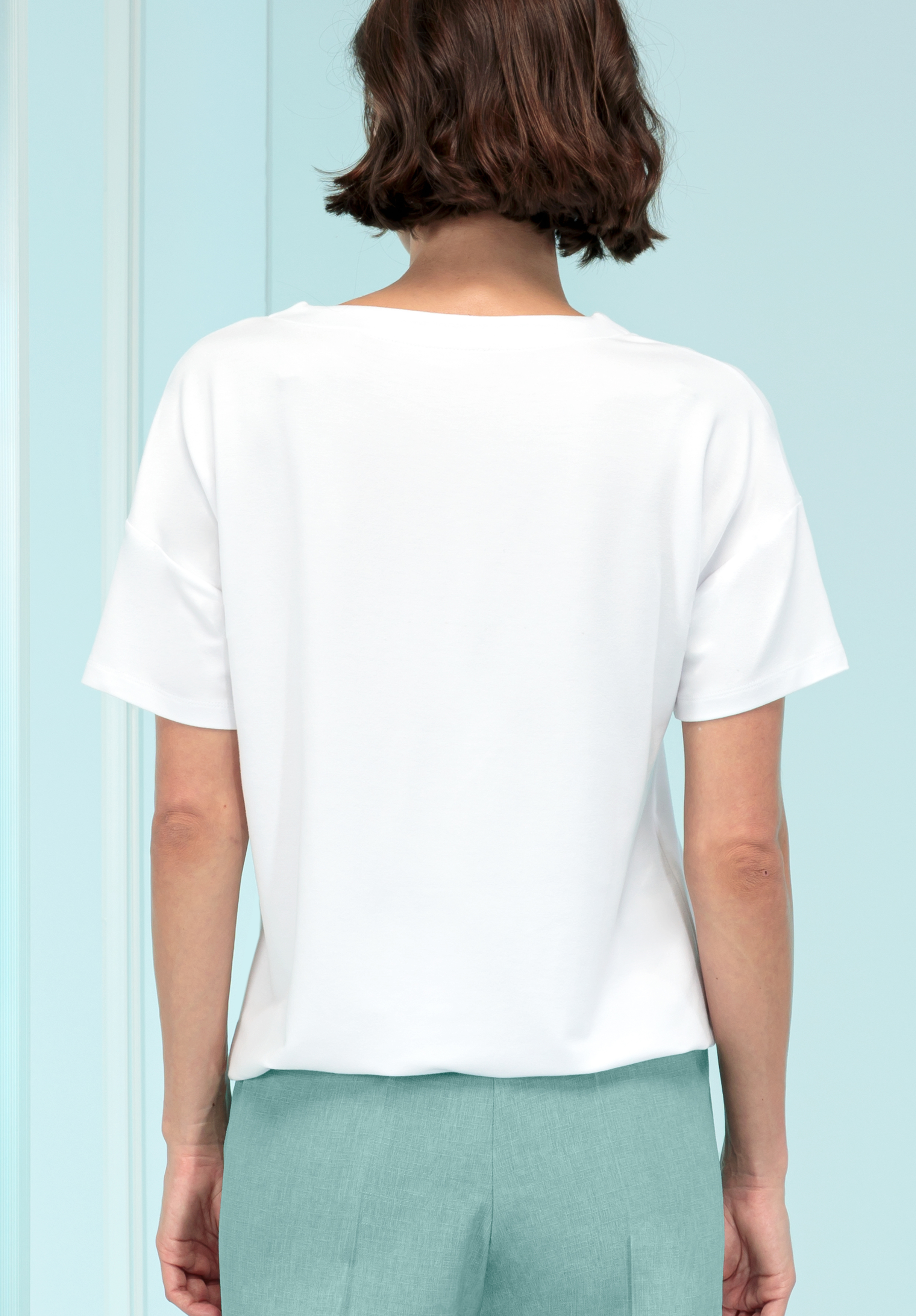 Von hinten ist eine Frau mit kurzen braunen Haaren zu sehen, die ein schlichtes weißes Kurzarm-T-Shirt und hellblau-grüne Hosen trägt und vor einem hellblauen Hintergrund steht.