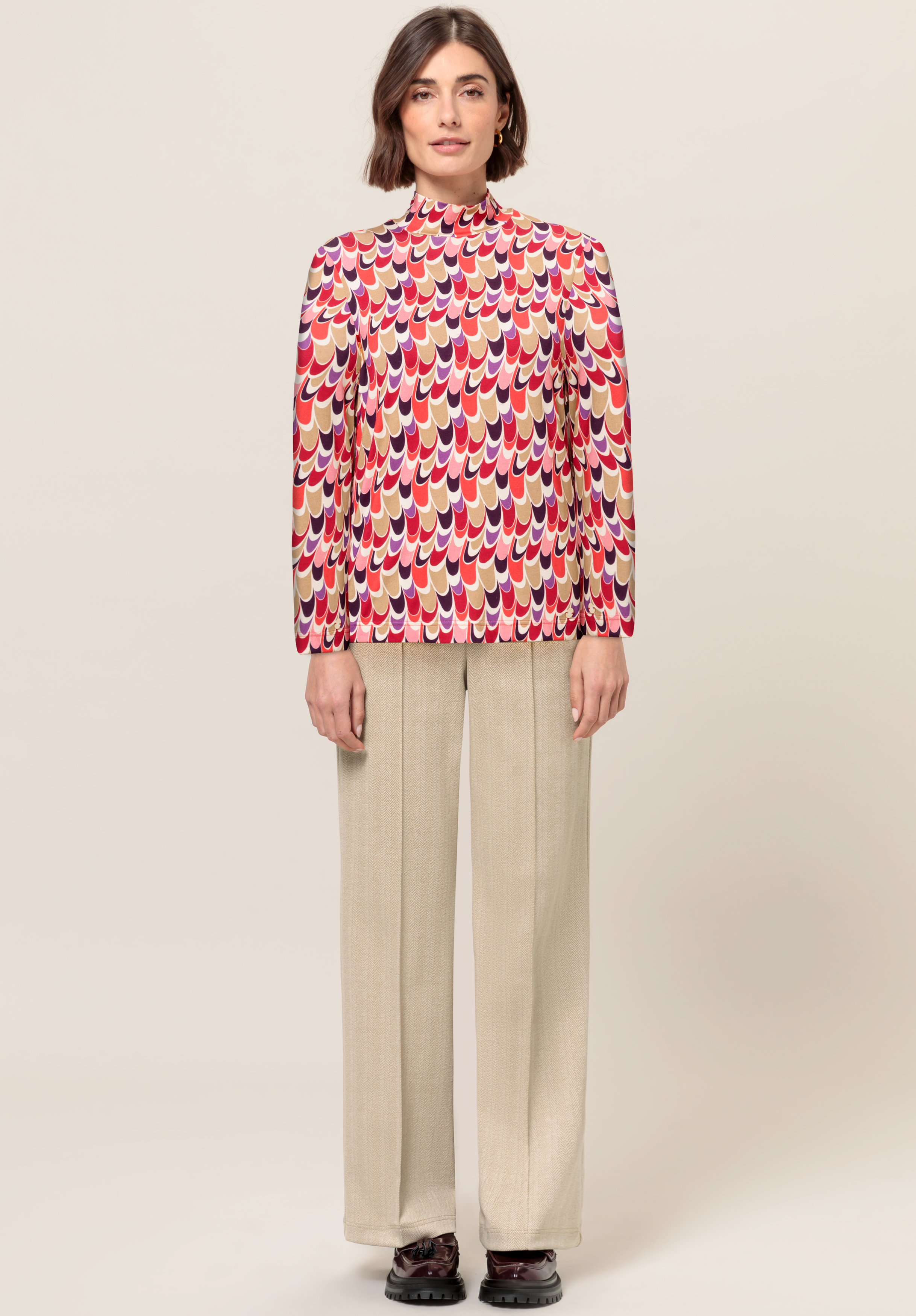 Eine Frau steht mit Blick nach vorne und trägt eine bunte Langarmbluse mit einem geometrischen Muster in Rot und Rosa, beige Hosen mit weitem Bein und dunkle Schuhe vor einem schlichten hellen Hintergrund.
