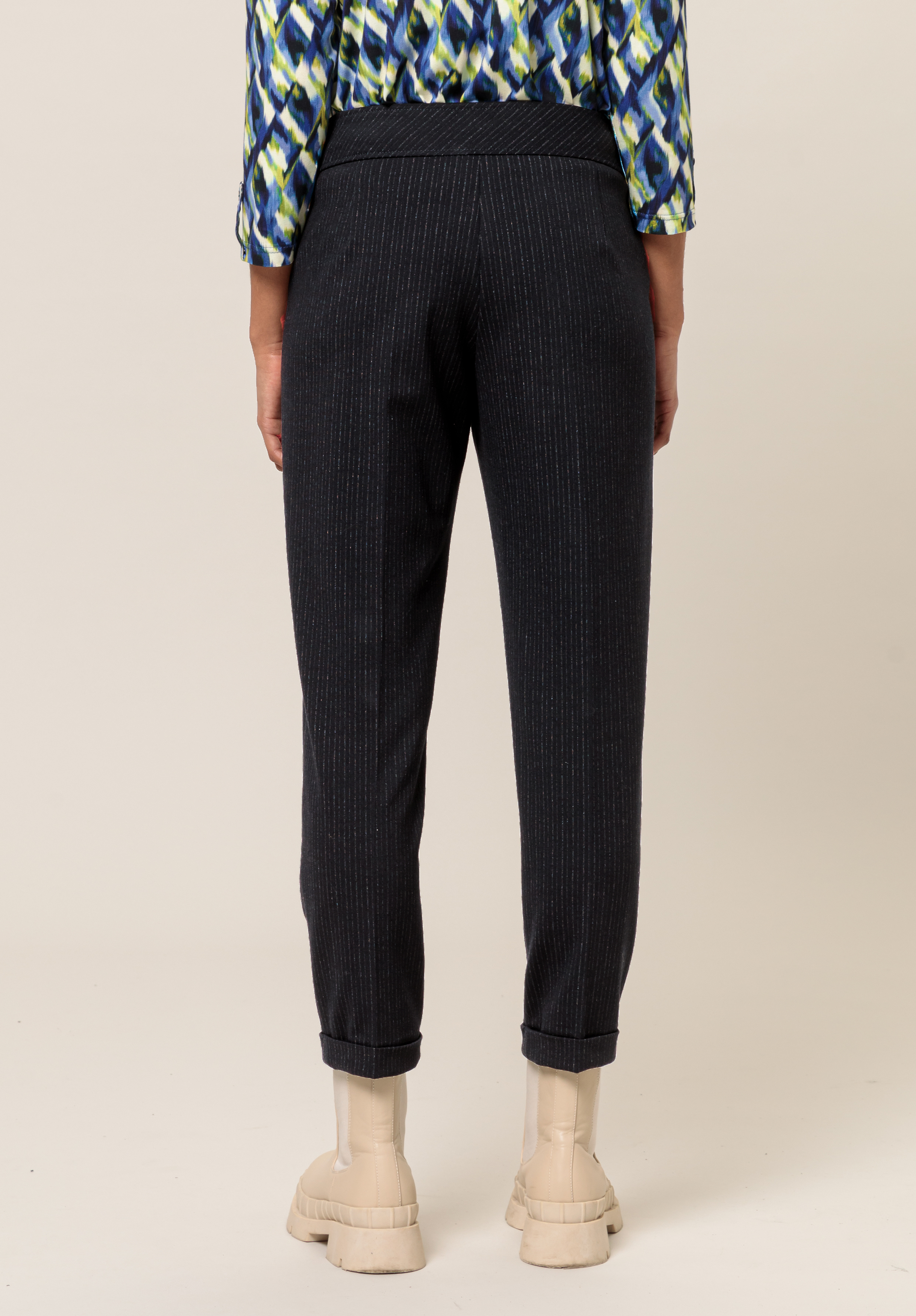 Eine Person steht mit abgewandtem Gesicht da und trägt eine schwarze Nadelstreifenhose, ein blaugrün gemustertes Oberteil und beige Stiefeletten. Der Hintergrund ist schlicht und neutral.