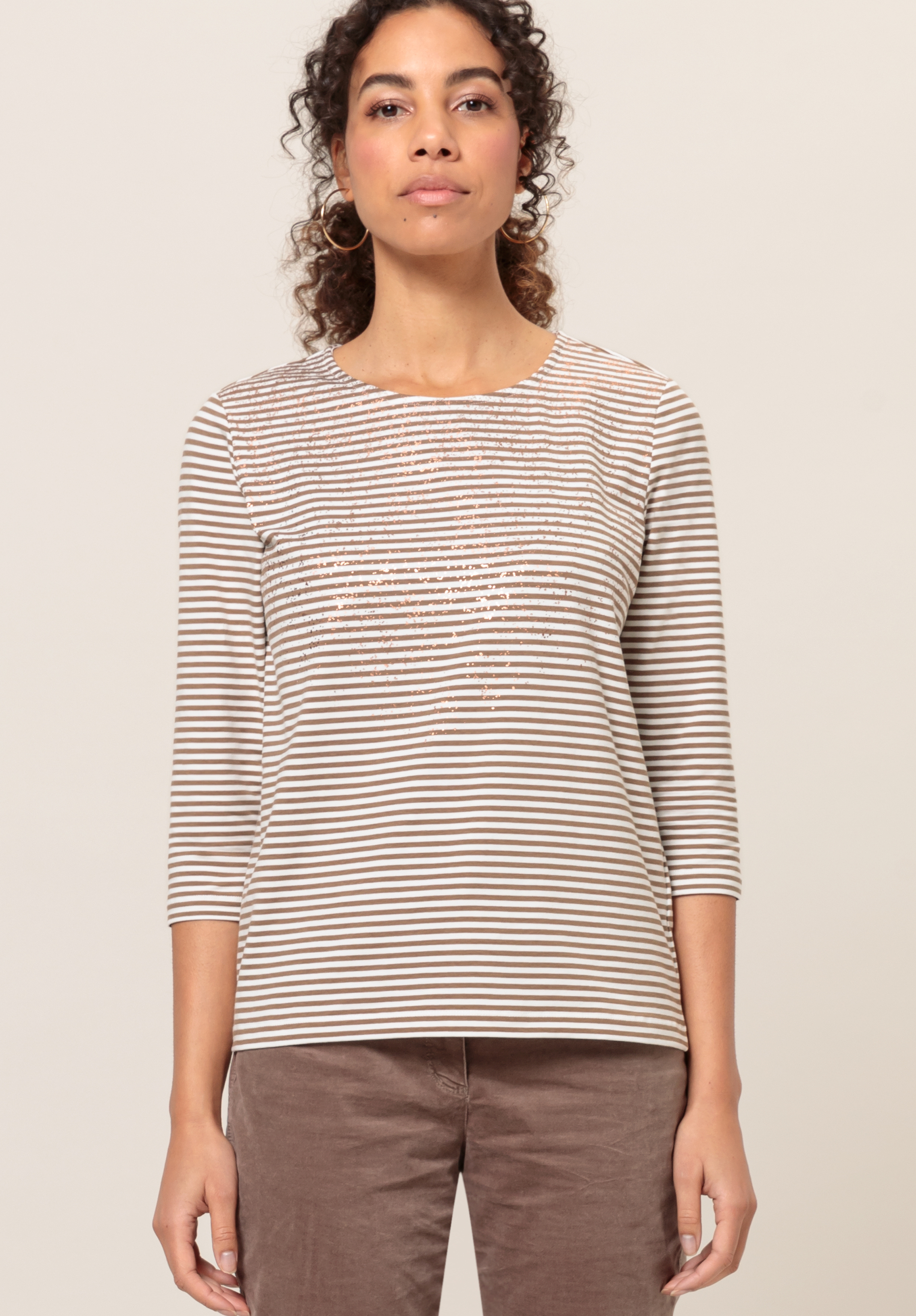 Rundhalsshirt DINI Eine Frau mit lockigem Haar, die ein beige-weiß gestreiftes Oberteil mit Dreiviertelärmeln und braune Hosen trägt, steht mit dem Gesicht nach vorne vor einem schlichten hellen Hintergrund.