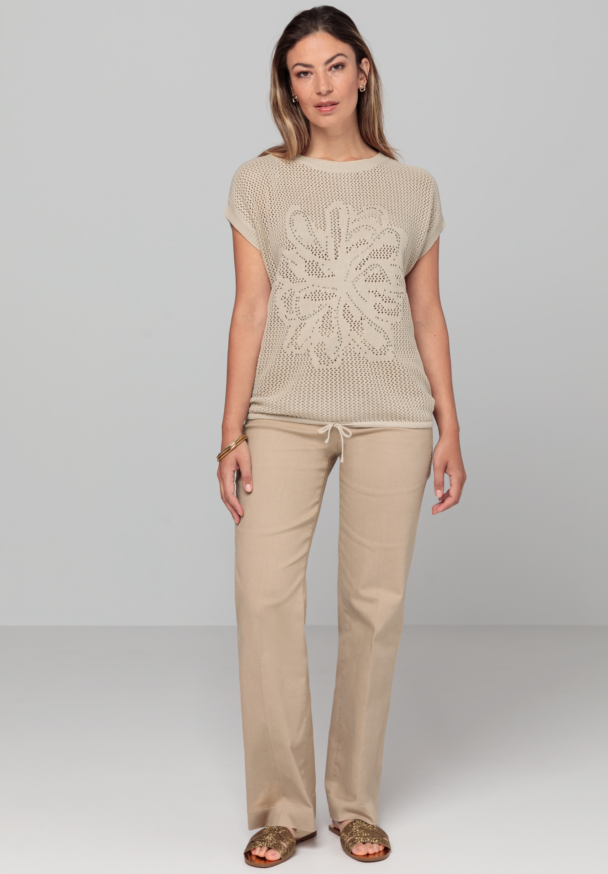Eine Frau steht mit dem Gesicht nach vorne. Sie trägt ein kurzärmeliges beigefarbenes Strickoberteil mit Blumenmuster, beigefarbene Hosen und goldene Sandalen. Der Hintergrund ist schlicht hellgrau.