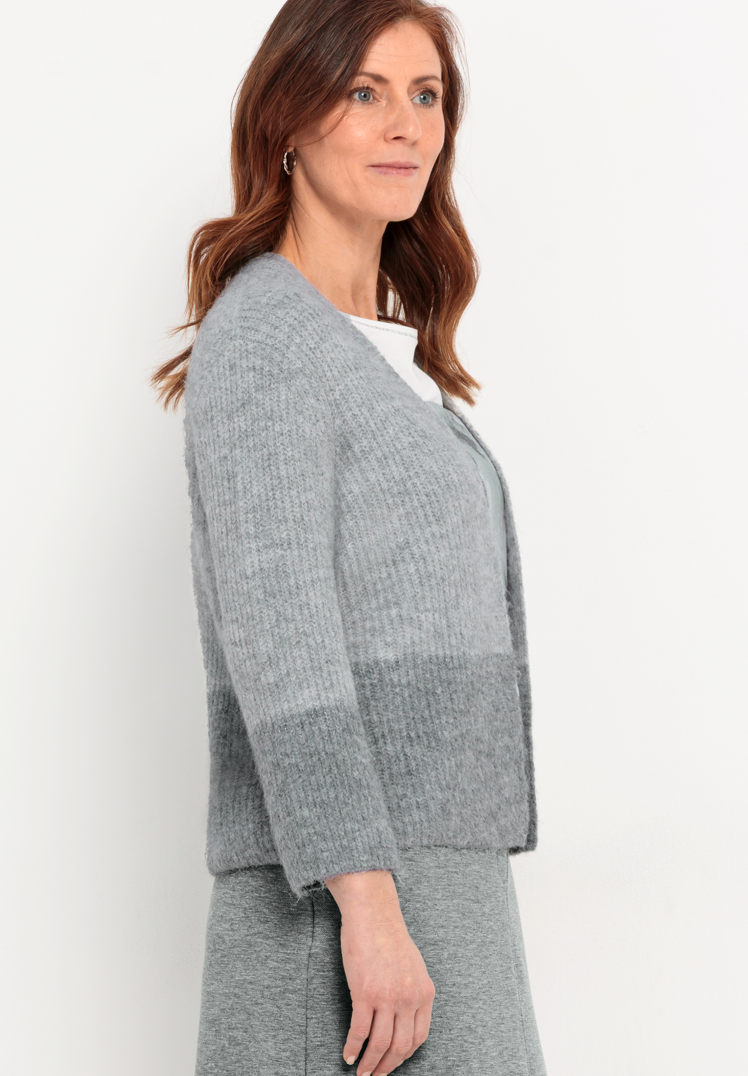Frau mit gewelltem braunem Haar, die eine hellgraue Strickjacke über einem grauen Kleid trägt, steht im Profil vor einem schlichten weißen Hintergrund.