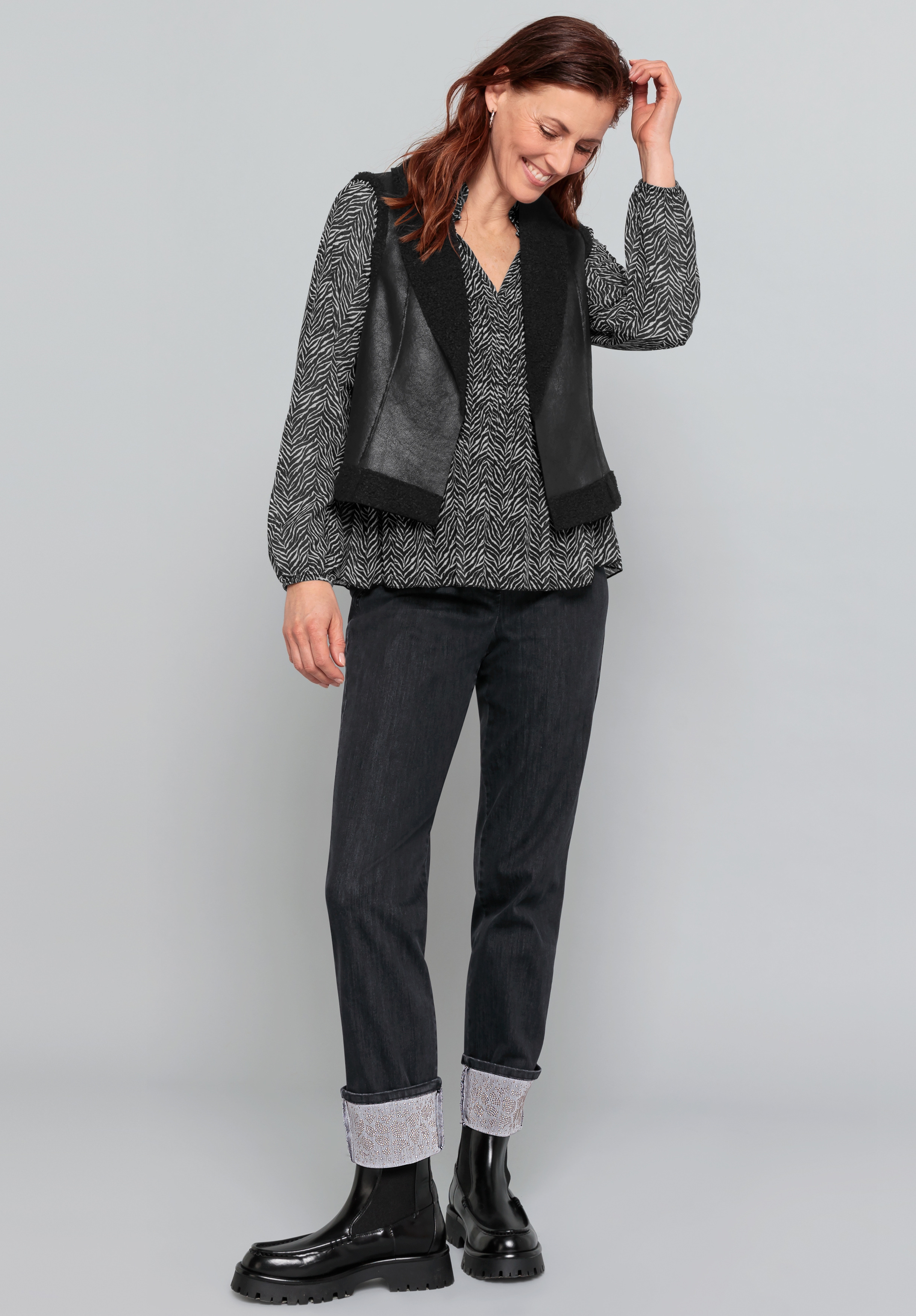Eine Frau lächelt und schaut nach unten, während sie ihr Haar streicht. Sie trägt eine gemusterte Bluse, eine schwarze Weste, eine dunkle Jeans mit Umschlag und glänzende schwarze Stiefel und steht vor einem schlichten hellgrauen Hintergrund.