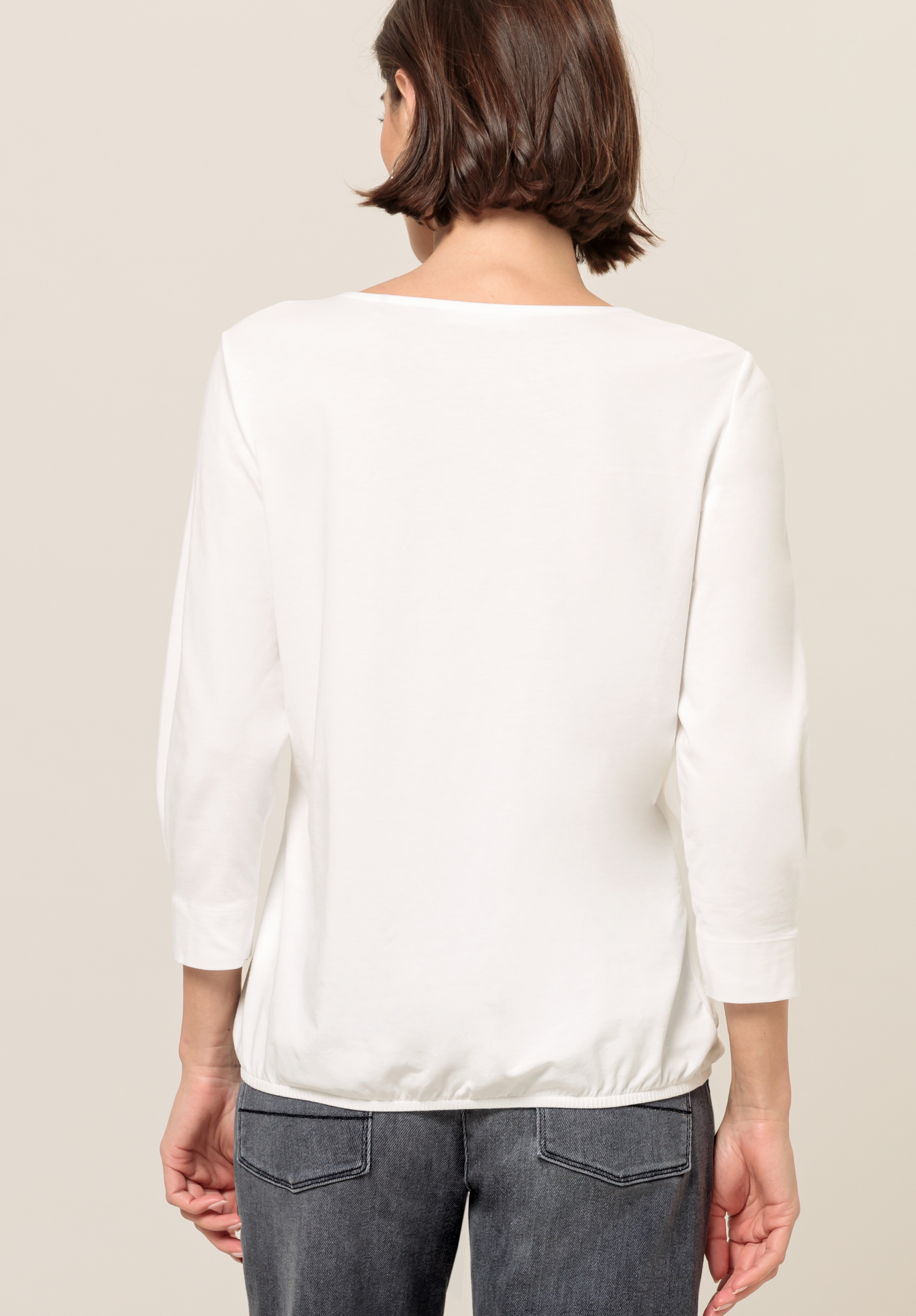 Eine Frau ist von hinten zu sehen. Sie trägt ein weißes, langärmliges Oberteil und dunkelgraue Jeans. Ihr Haar ist kinnlang und braun. Der Hintergrund ist schlicht und hell.