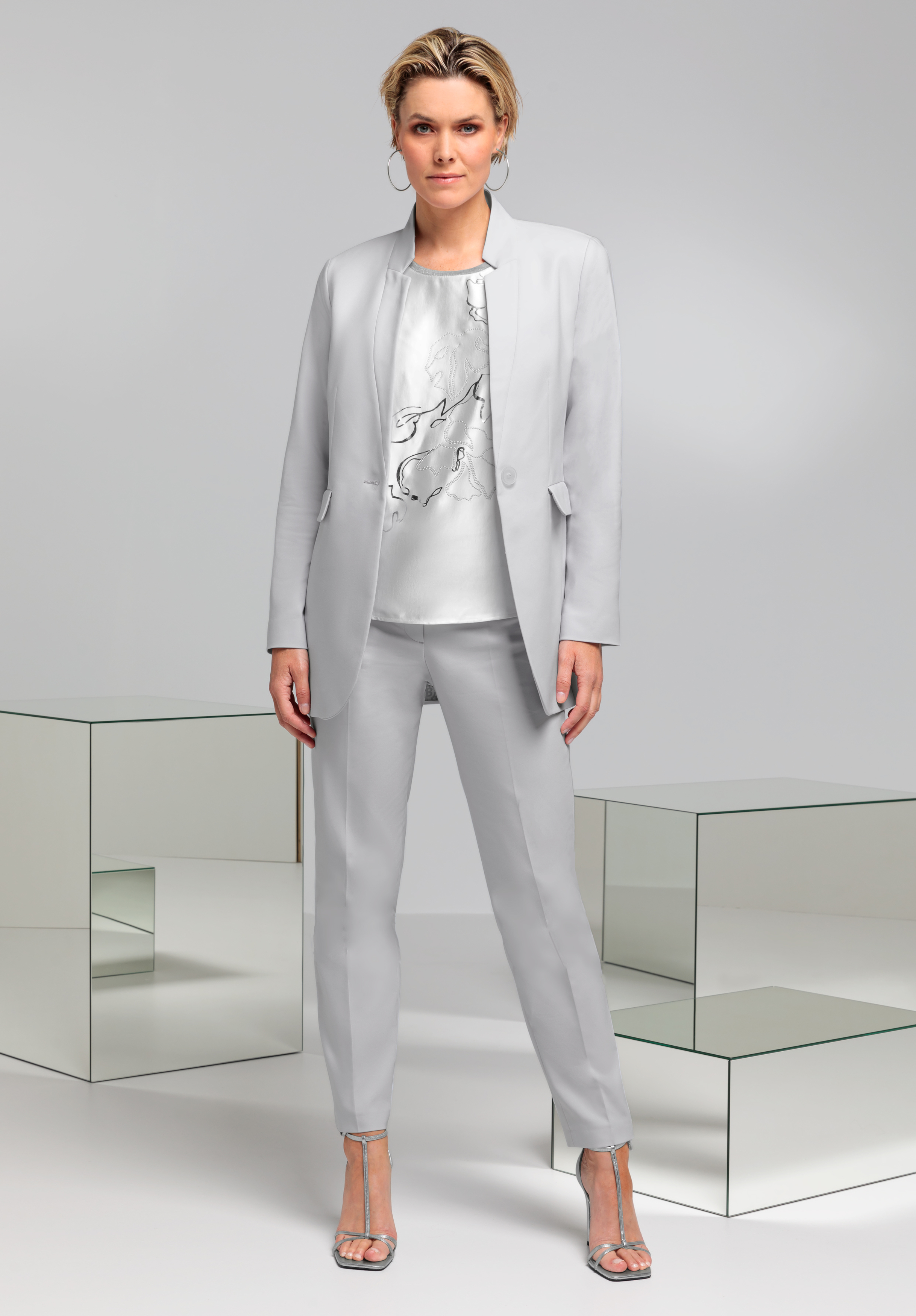 Eine Frau steht selbstbewusst in einem hellgrauen Hosenanzug und passenden Heels. Unter ihrem Blazer trägt sie eine gemusterte Bluse. Der Hintergrund ist hell, neben ihr spiegelnde geometrische Würfel.