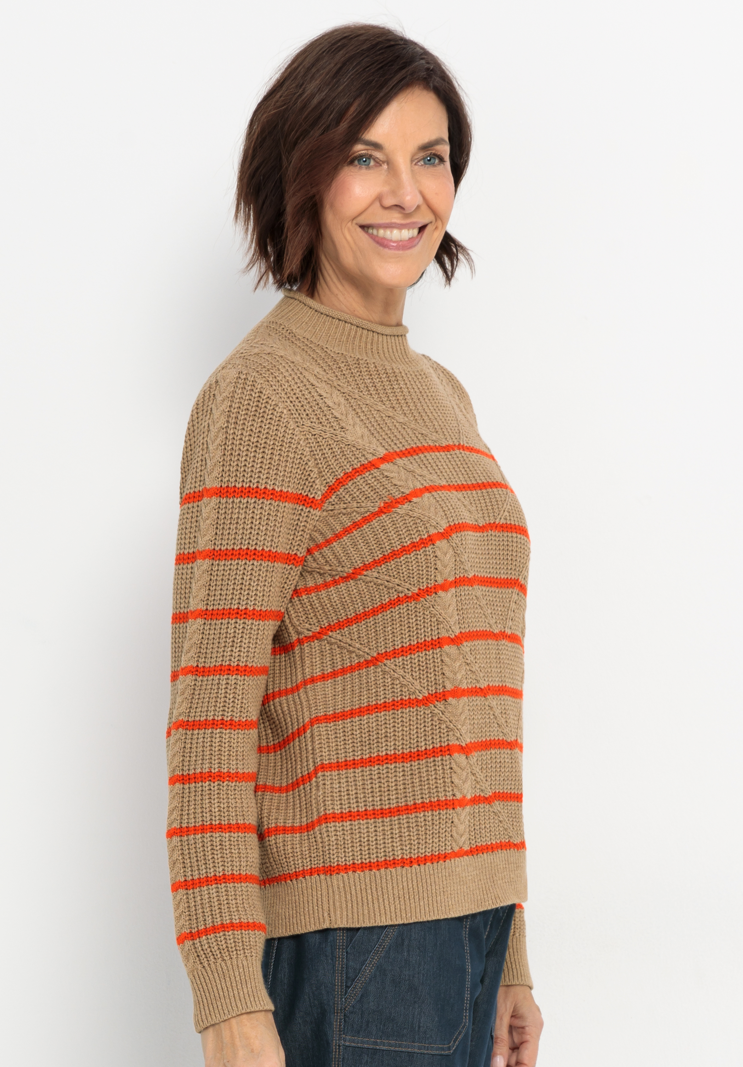 Eine Frau mit kurzen braunen Haaren lächelt, während sie einen hellbraunen Pullover mit roten Querstreifen und eine dunkelblaue Hose trägt und vor einem schlichten weißen Hintergrund steht.