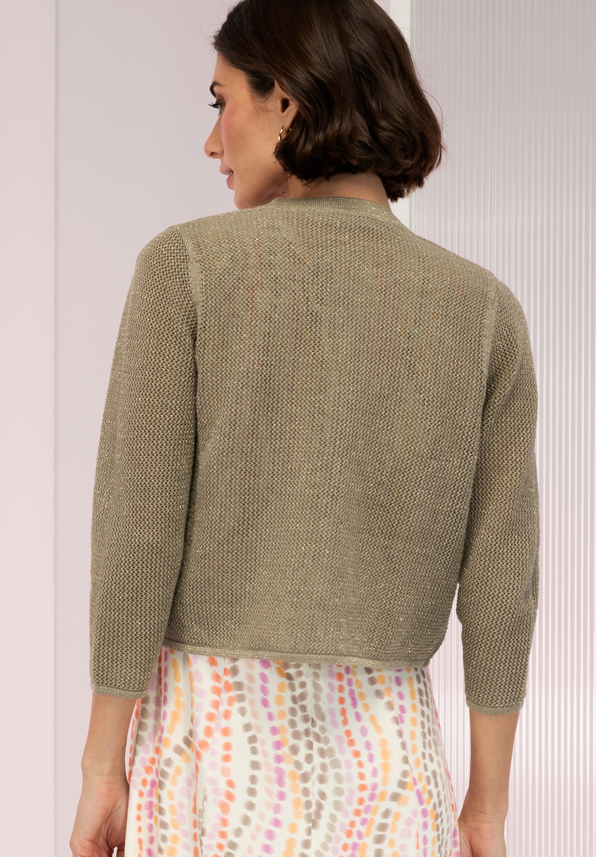 Von hinten ist eine Frau mit kurzen braunen Haaren zu sehen, die eine strukturierte, hellbraune Strickjacke mit Dreiviertelärmeln über einem weißen Kleid mit einem bunten Punktmuster trägt.