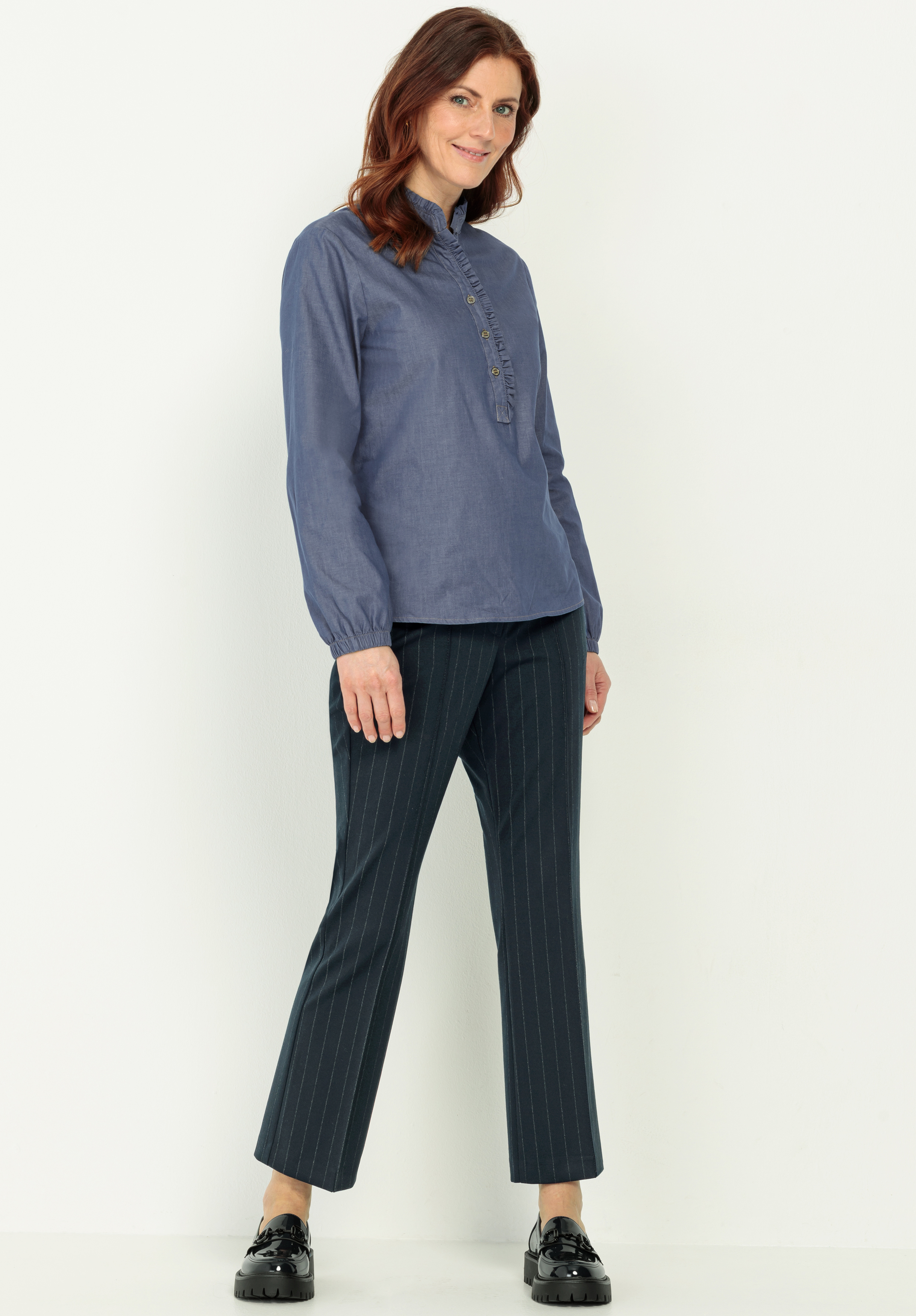 Eine Frau steht vor einem schlichten, hellen Hintergrund. Sie trägt ein langärmeliges blaues Button-up-Hemd und eine dunkelblaue Nadelstreifenhose mit schwarzen, glänzenden Schuhen. Sie lächelt und blickt leicht zur Seite.