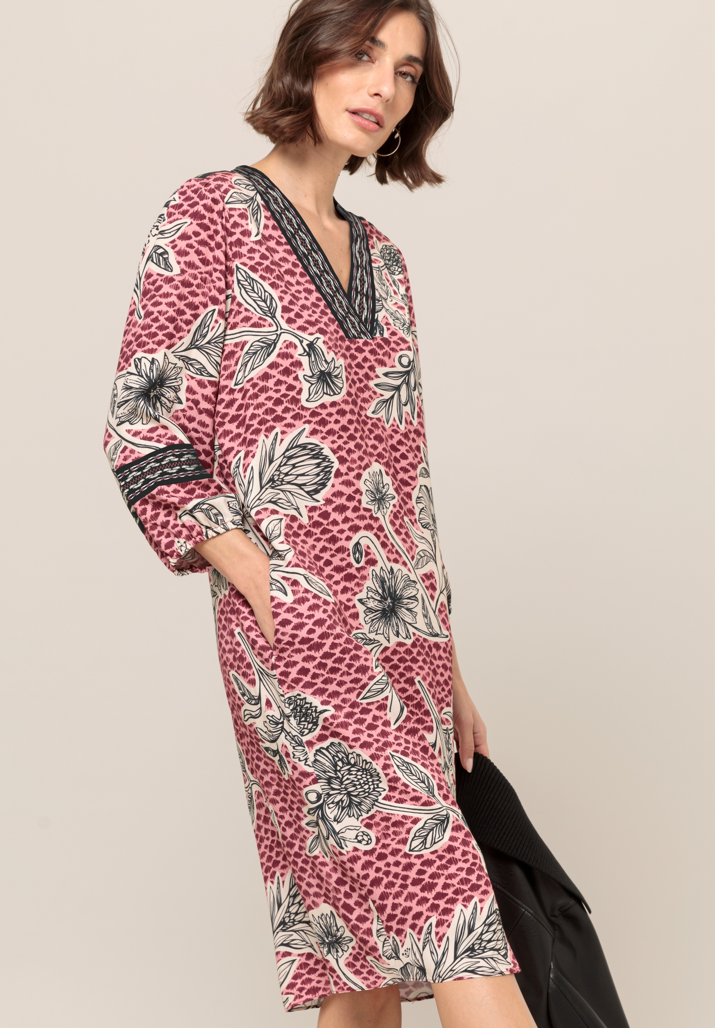 Eine Frau trägt ein locker sitzendes, knielanges Kleid mit rosa-weißem Blumenmuster und schwarzem Besatz an Ausschnitt und Ärmelbündchen. Sie hat kurze braune Haare und hält einen dunklen Mantel über dem Arm.