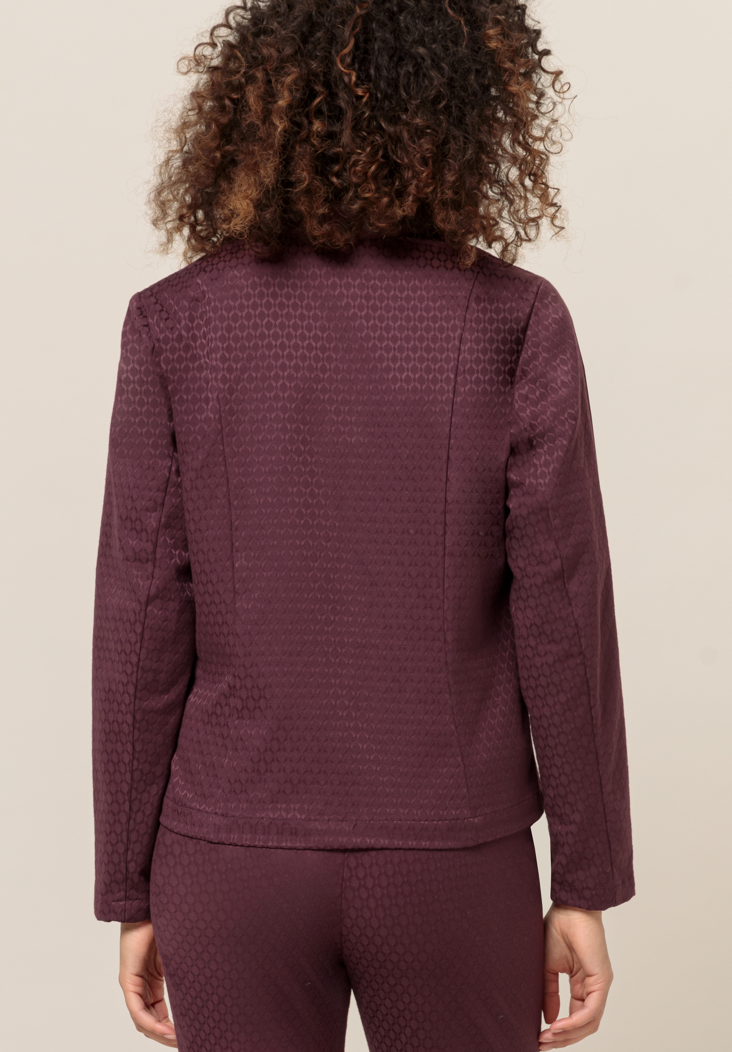 Eine Person mit lockigem Haar, die eine strukturierte, dunkelviolette Jacke und eine dazu passende Hose trägt, von hinten vor einem schlichten hellen Hintergrund gezeigt.