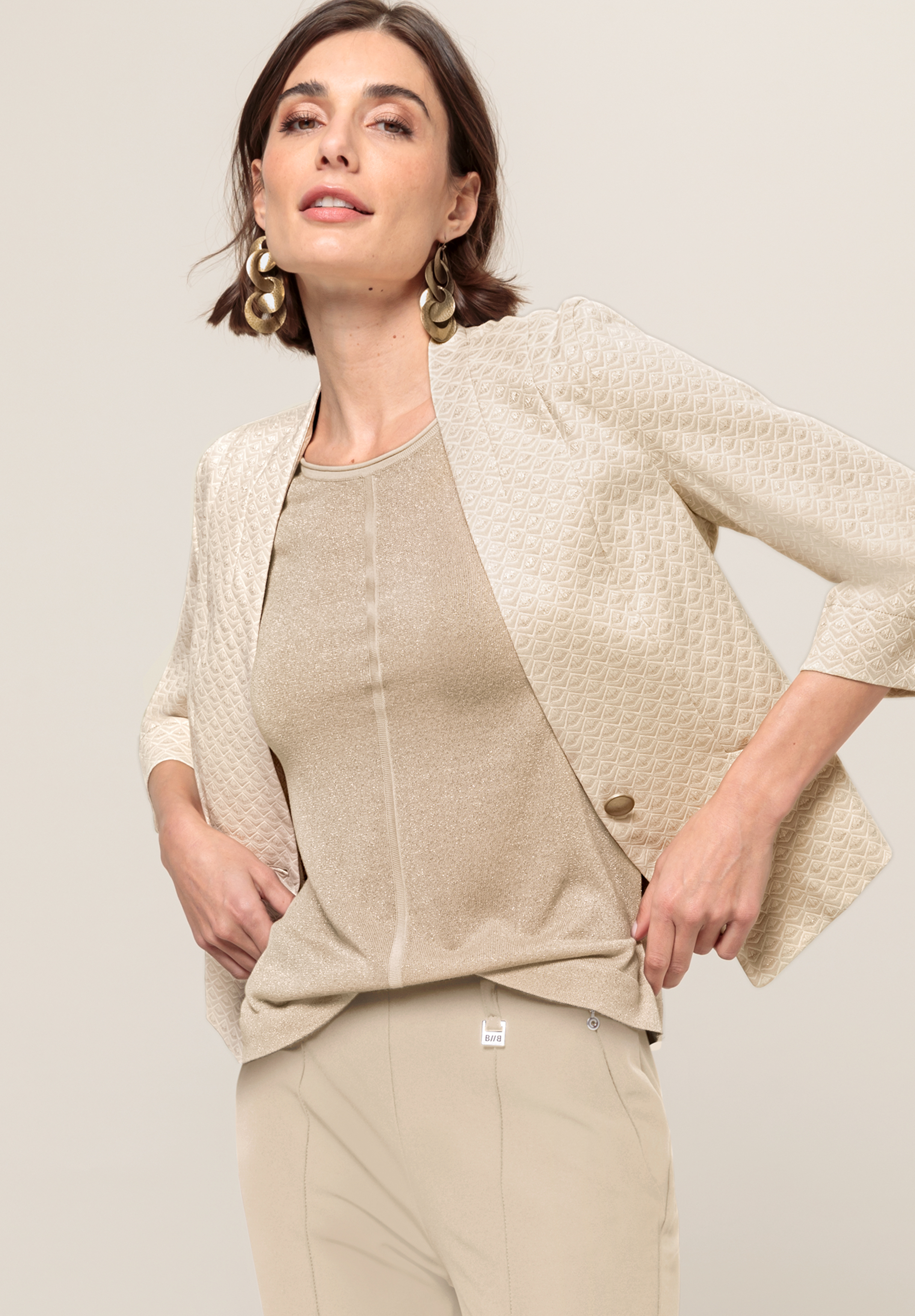 Kurzblazer ABBY Eine Frau in einem strukturierten beigen Blazer, einem schimmernden beigen Oberteil und einer hellbeigen Hose posiert selbstbewusst mit den Händen in den Hüften vor einem schlichten Hintergrund. Sie hat kurze braune Haare und trägt baumelnde Ohrringe.