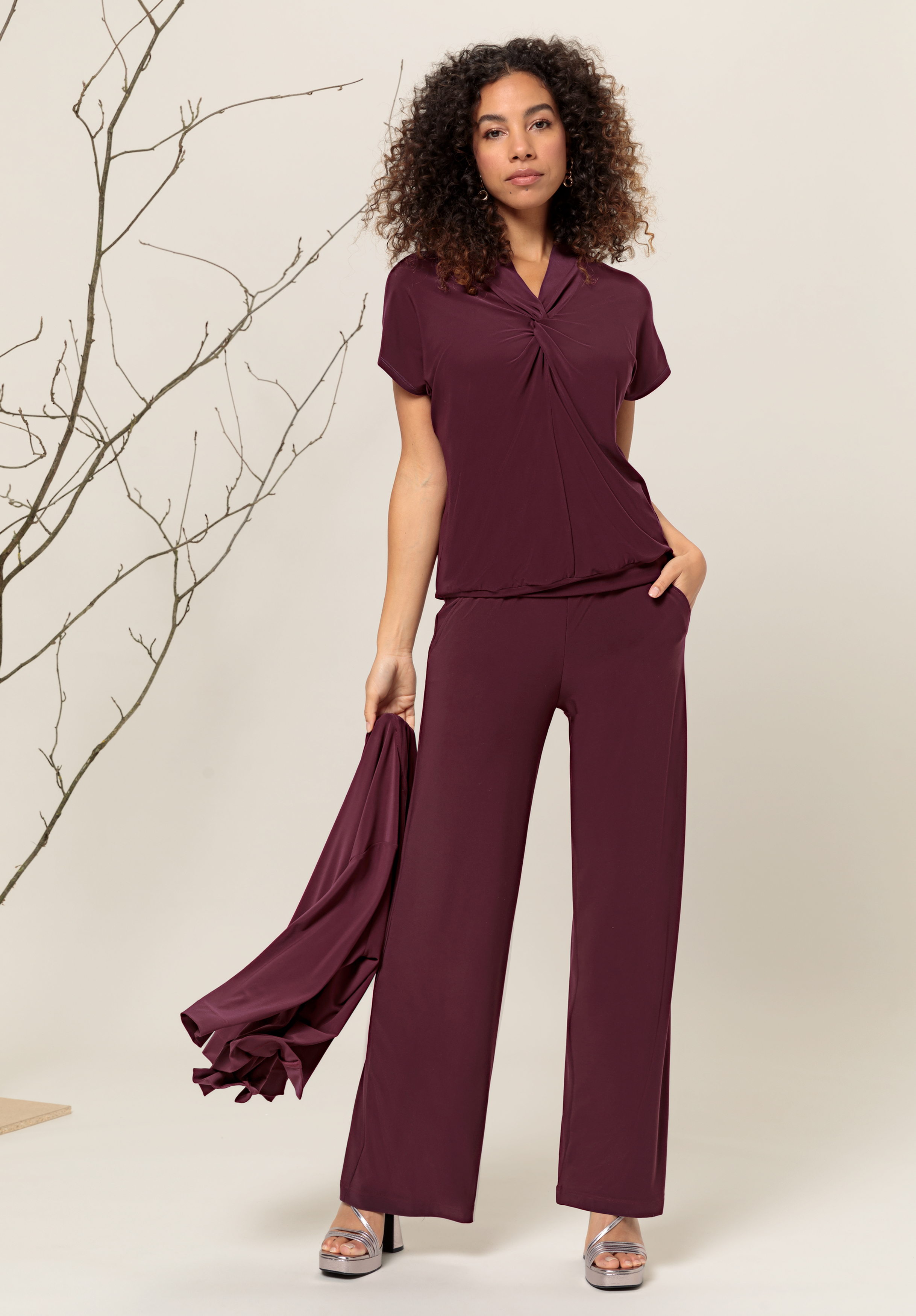 Eine Frau mit lockigem Haar trägt ein burgunderfarbenes Kurzarmoberteil und eine dazu passende Hose mit weitem Bein. Sie hält eine passende Jacke in der Hand. Sie steht mit einer Hand in der Tasche und posiert neben einem kahlen, blattlosen Ast.