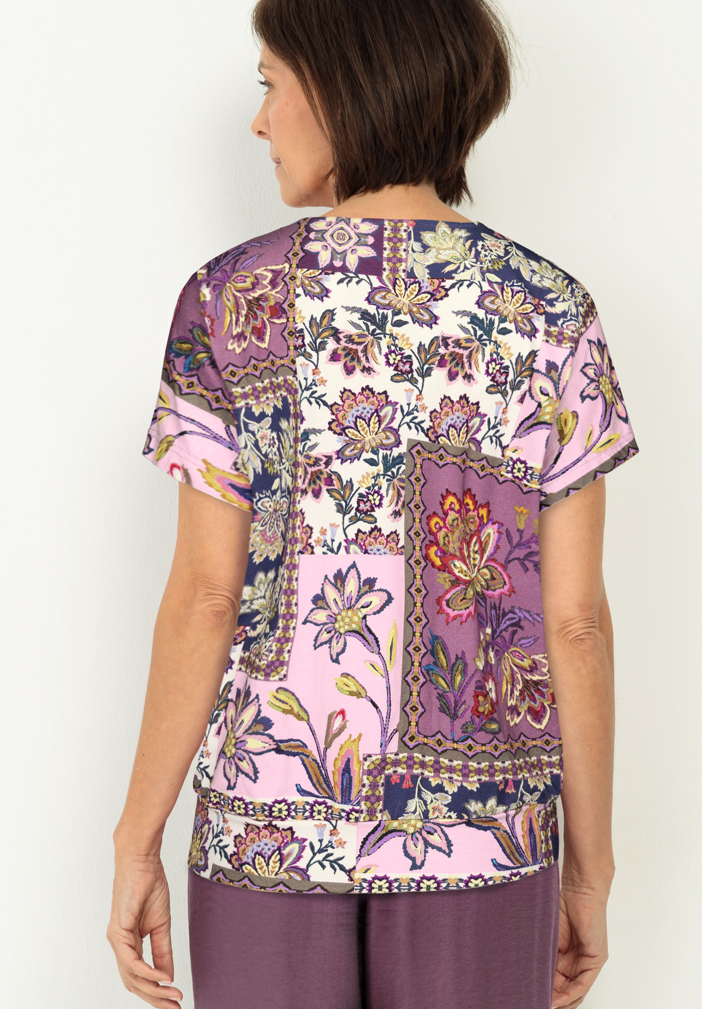 Eine Frau mit kurzen braunen Haaren steht mit dem Rücken zur Kamera. Sie trägt eine kurzärmelige Bluse mit einem rosa, lila und cremefarbenen Blumenmuster und dazu eine lila Hose.