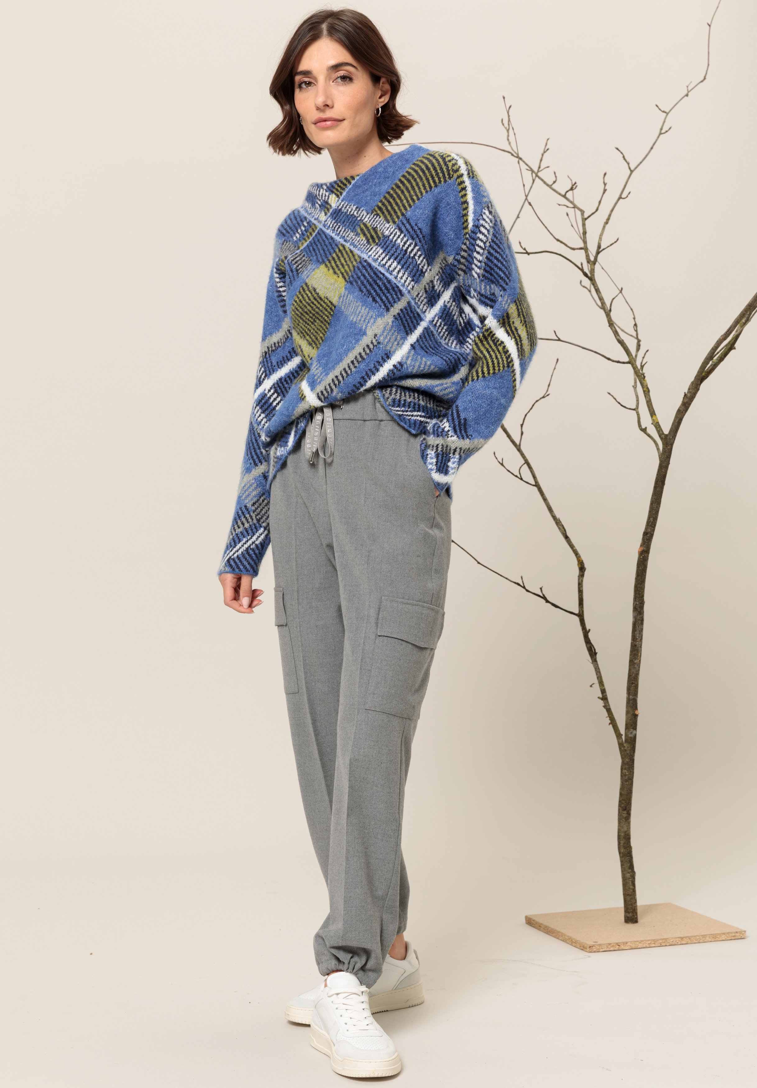 Eine Frau steht in einem Haus und trägt einen blau-grün-weiß karierten Pullover, hellgraue Cargohosen mit Kordelzug und weiße Turnschuhe. Sie posiert neben einem minimalistischen kahlen Ast vor beigem Hintergrund.