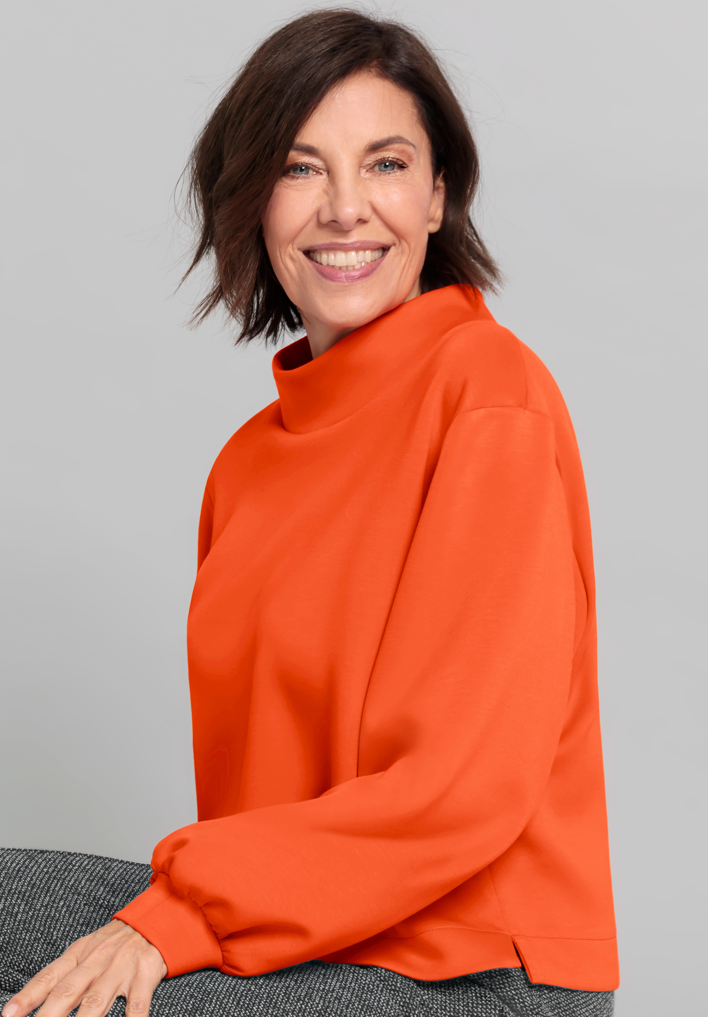 Eine lächelnde Frau mit kurzen braunen Haaren trägt einen orangefarbenen Rollkragenpullover und eine graue Hose und sitzt vor einem schlichten hellgrauen Hintergrund.