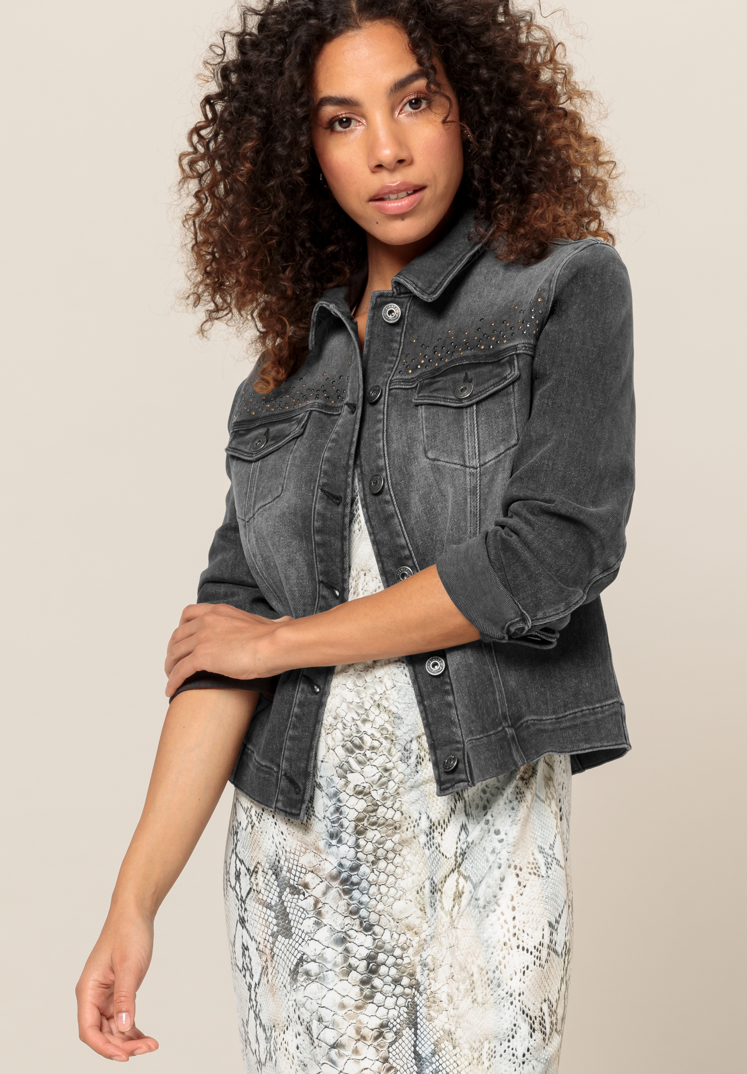 Jeansjacke JUNE Eine Frau mit lockigem Haar trägt eine schwarze Jeansjacke über einem hellen Kleid mit Schlangenmuster und steht vor einem schlichten beigen Hintergrund.